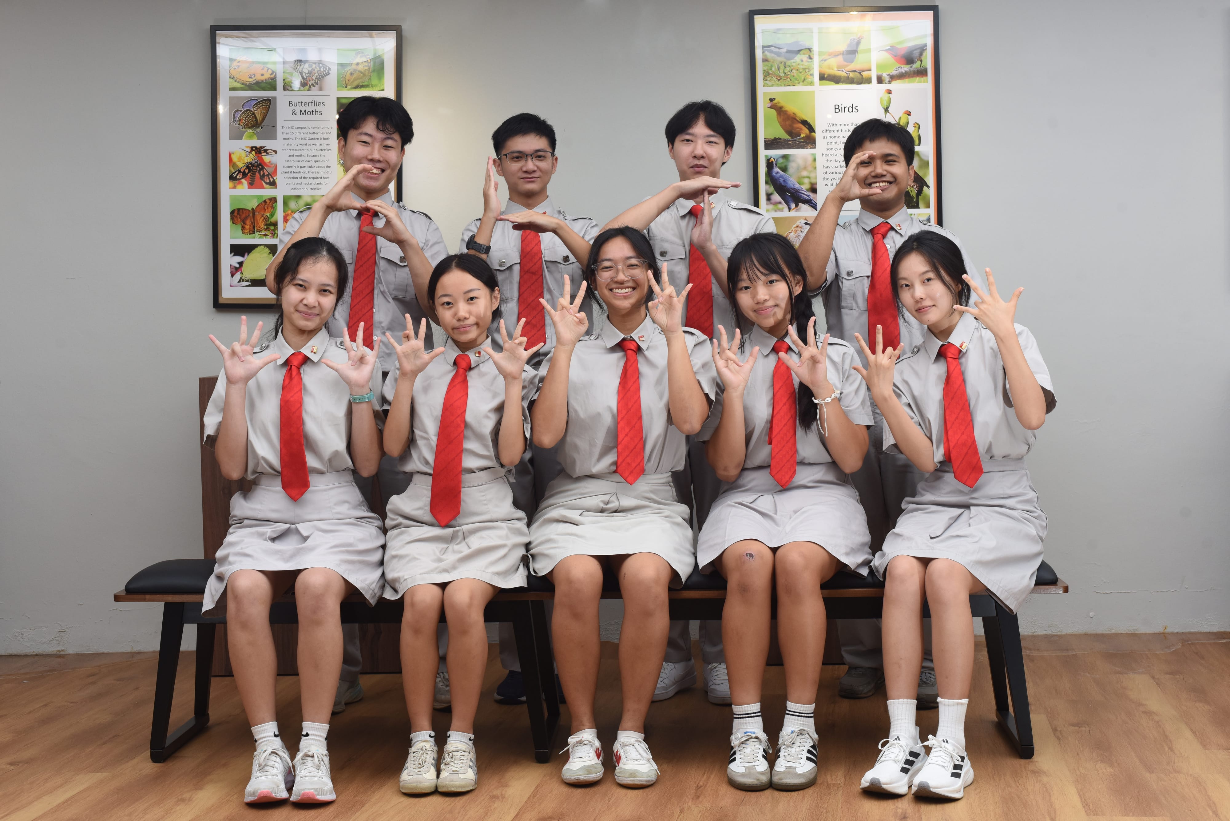 Nine students in gray uniforms with red ties pose in two rows, holding up fingers, in front of nature posters.