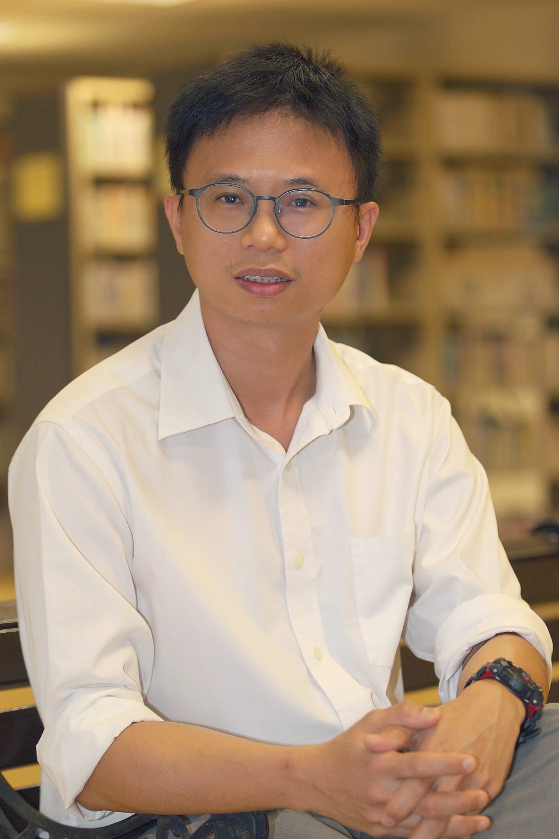A man with glasses and short dark hair wearing a white shirt sits with hands clasped, bookshelves blurred behind him.