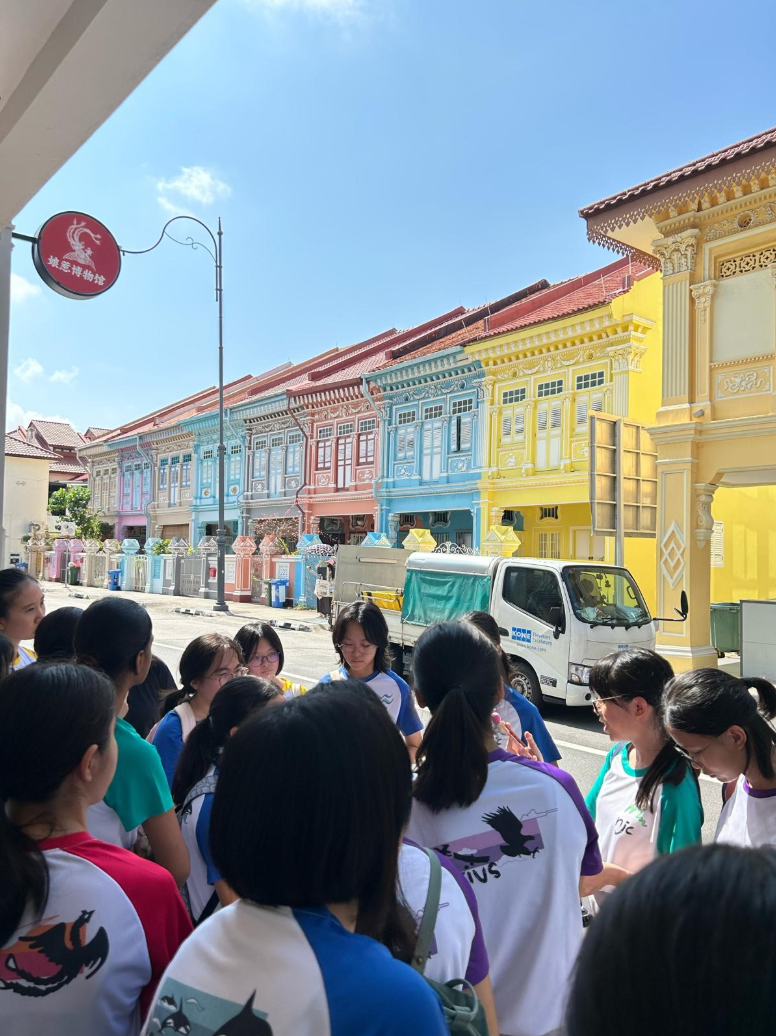 A group of students stands on a street facing colorful Peranakan shophouses in pastel blue, pink, and yellow.