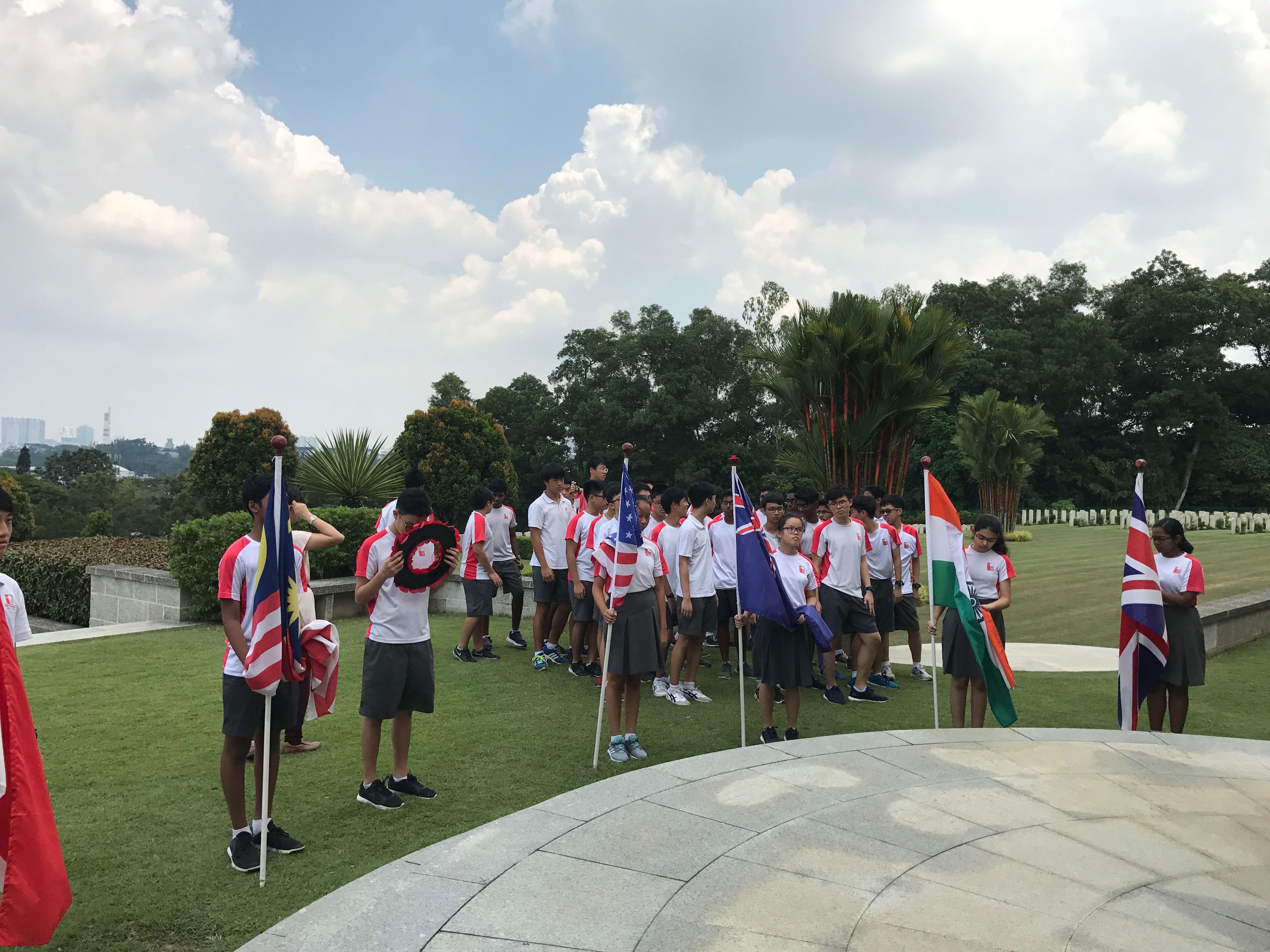 Students in white and red uniforms hold flags of Malaysia, Australia, India, and UK at an outdoor memorial ceremony.