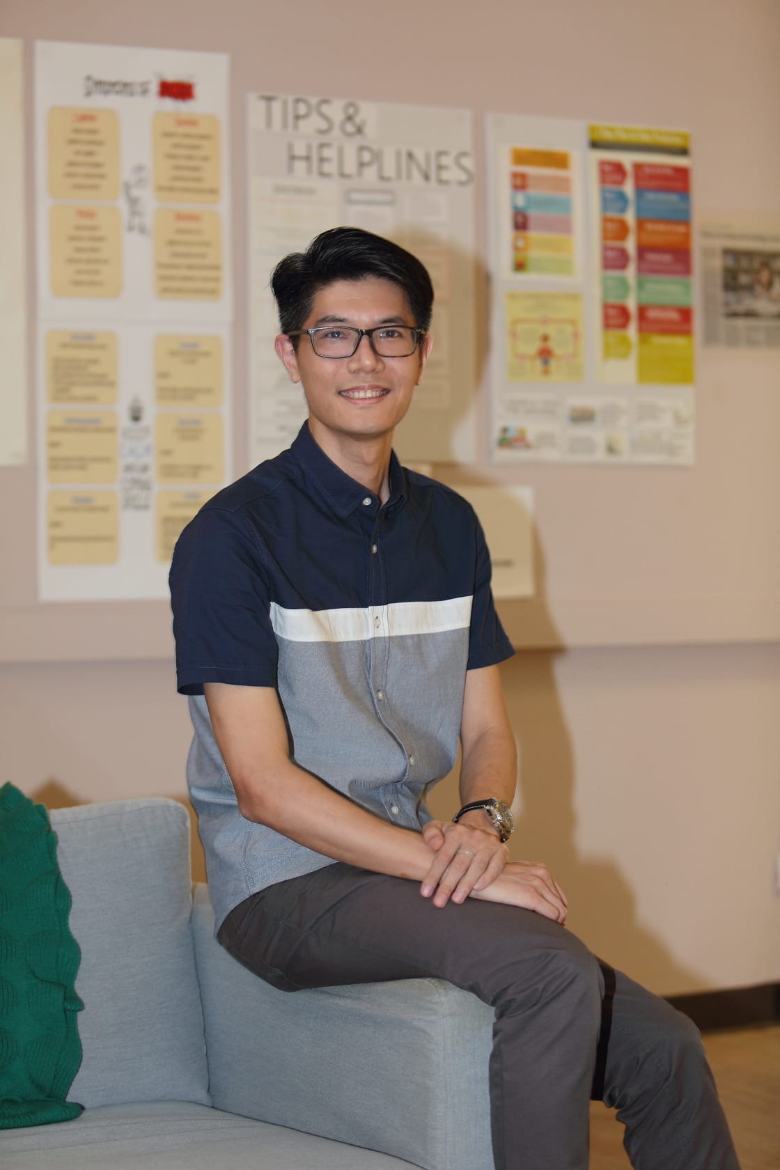 A young man with glasses sits smiling on a gray sofa, with a Tips and Helplines poster on the wall behind him.