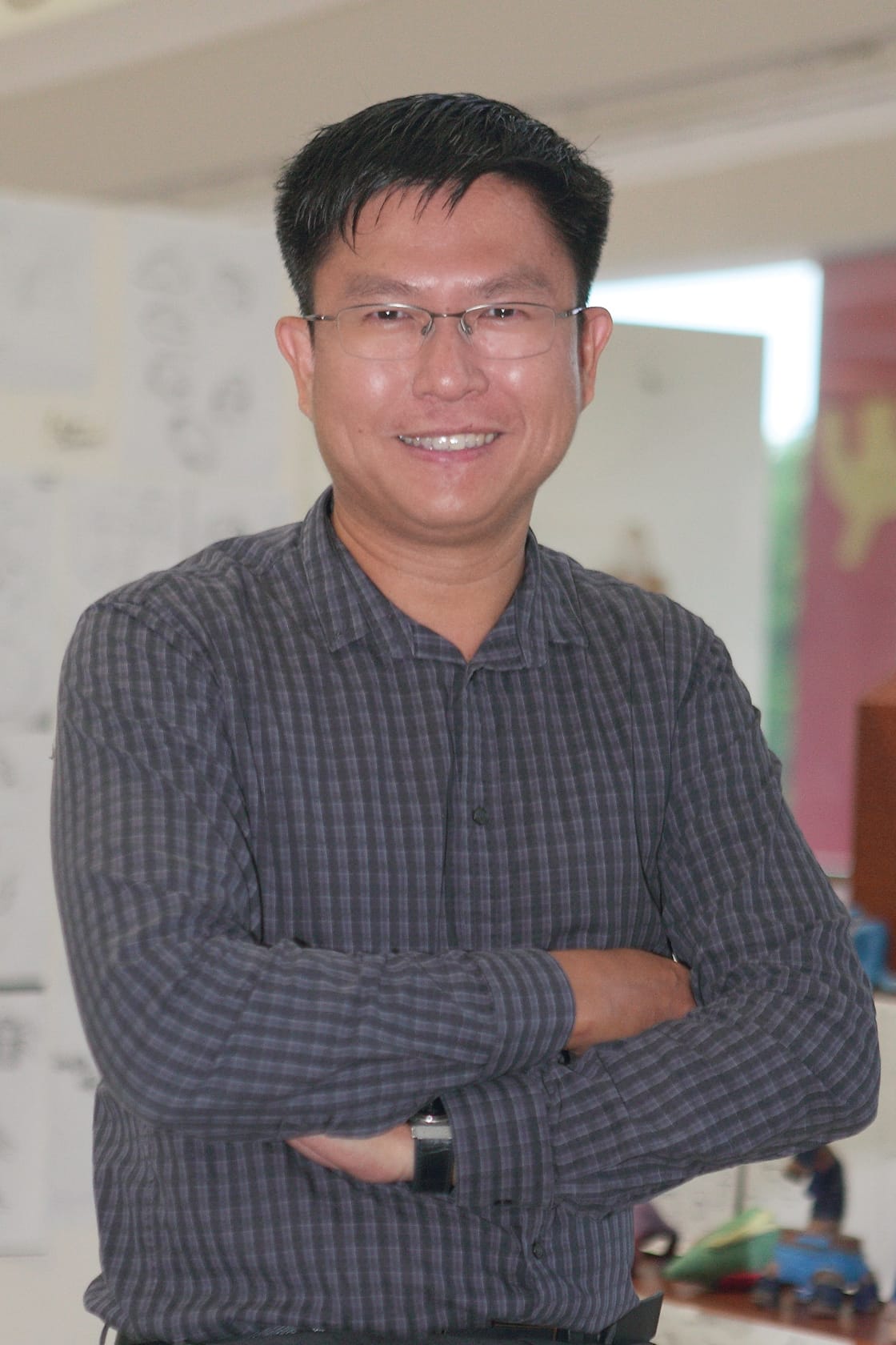 A smiling man with glasses and crossed arms wearing a gray plaid shirt, standing in a blurred indoor setting.