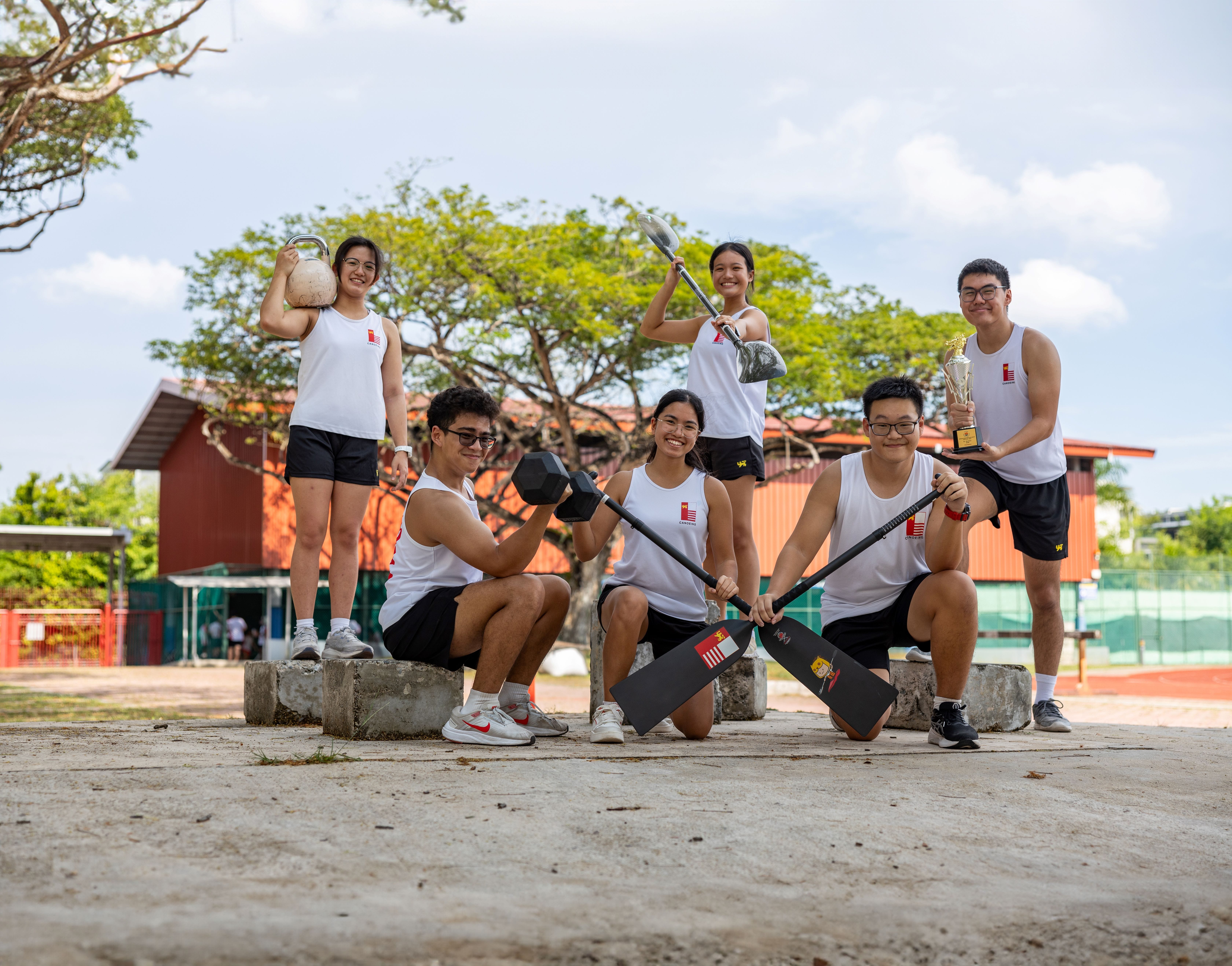 Six young athletes in white tank tops pose outdoors holding paddles, a kettlebell, dumbbell, and a trophy.