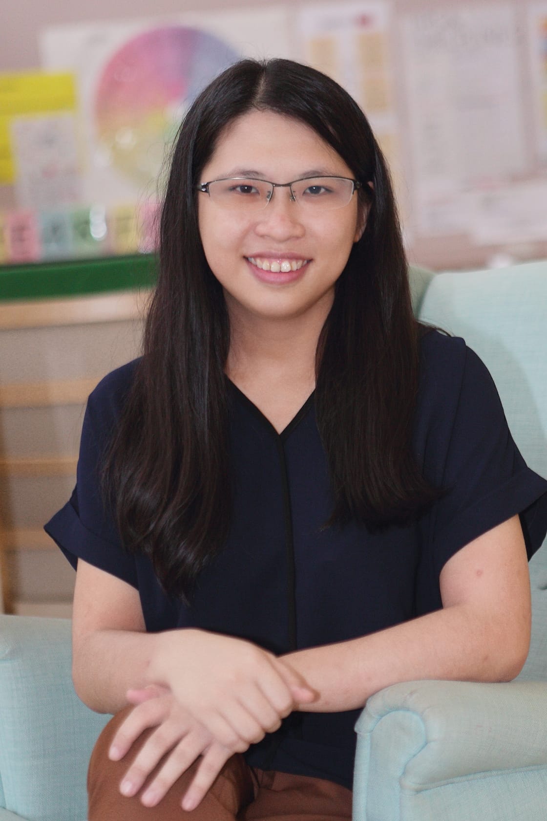 A smiling woman with long black hair and glasses, wearing a navy top, seated on a light blue chair with hands folded.