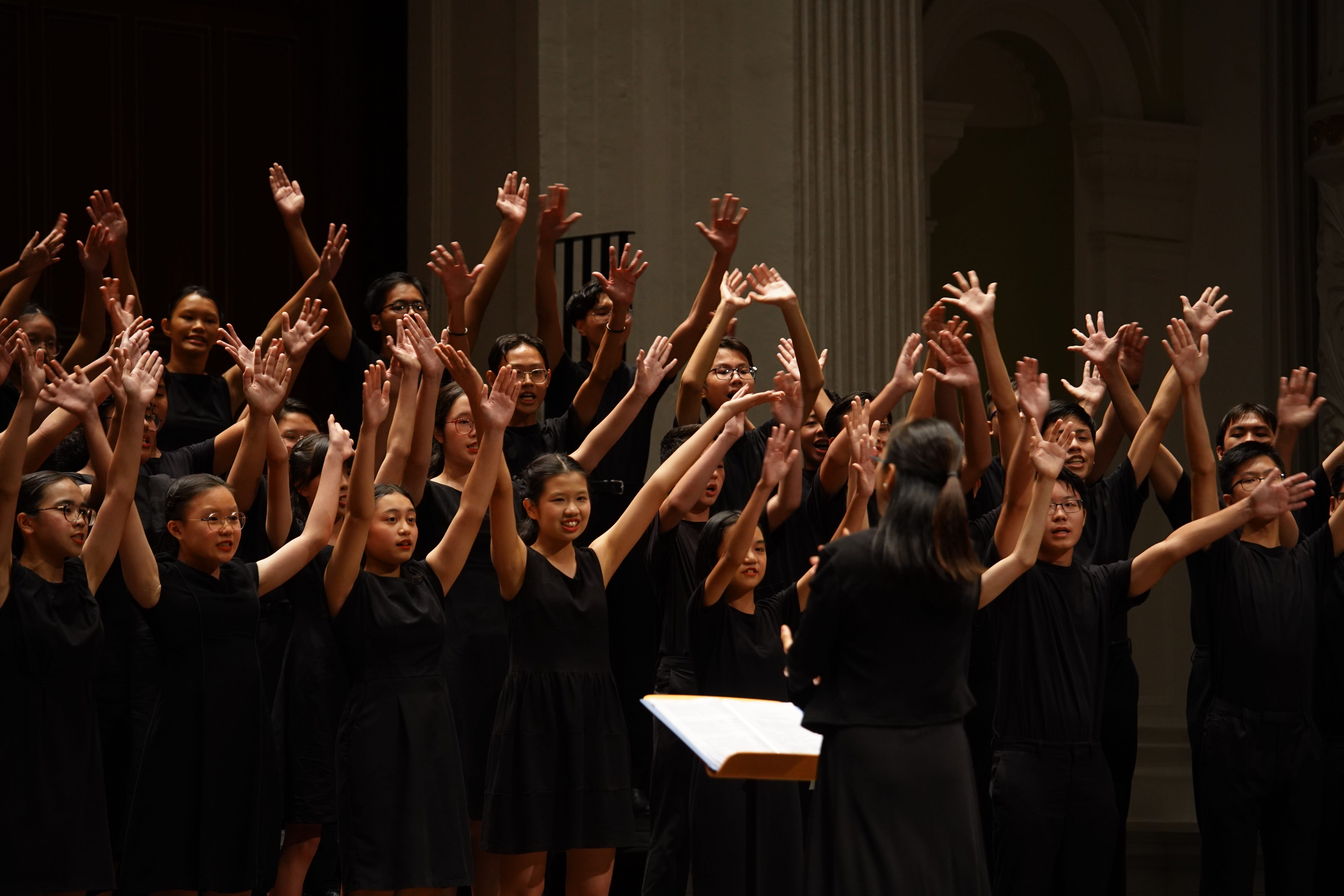 A large youth choir in black outfits raises their hands on stage while a conductor stands before a music stand.