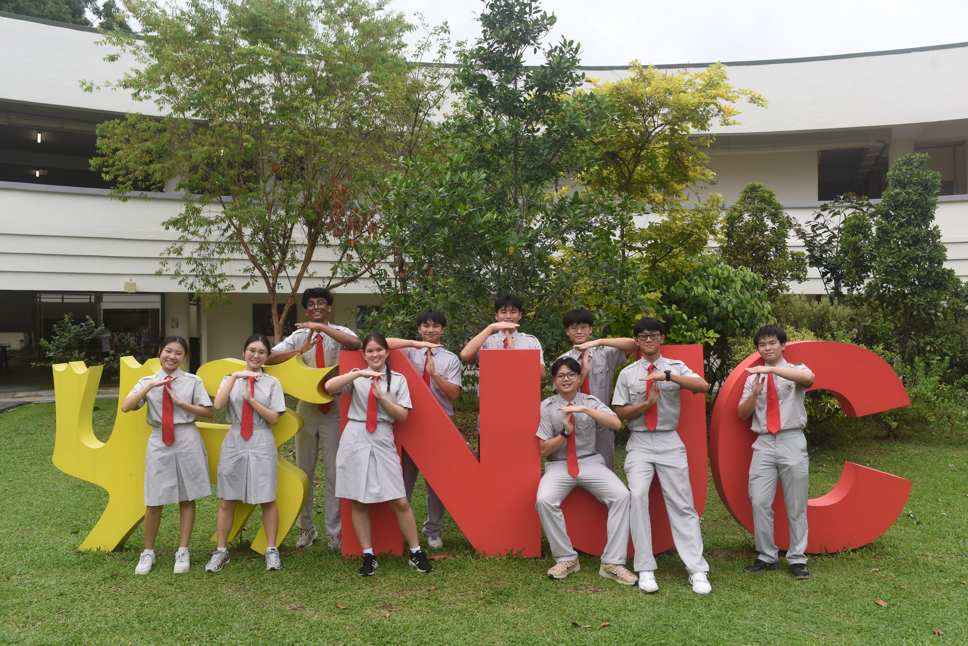 Nine students in gray uniforms pose by large red and yellow letters on a grassy courtyard with trees