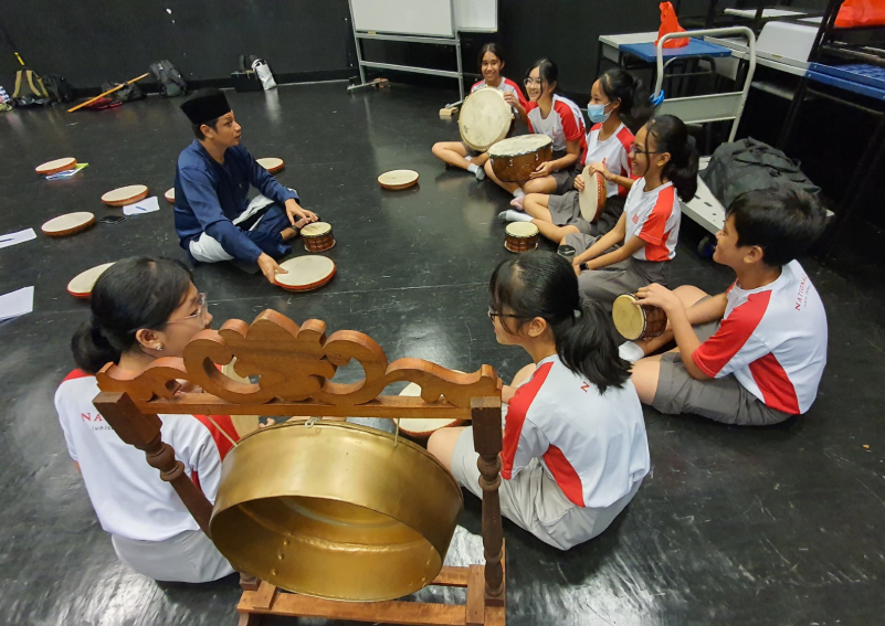 Students in uniforms sit in a circle on a dark floor learning traditional percussion instruments with an instructor.