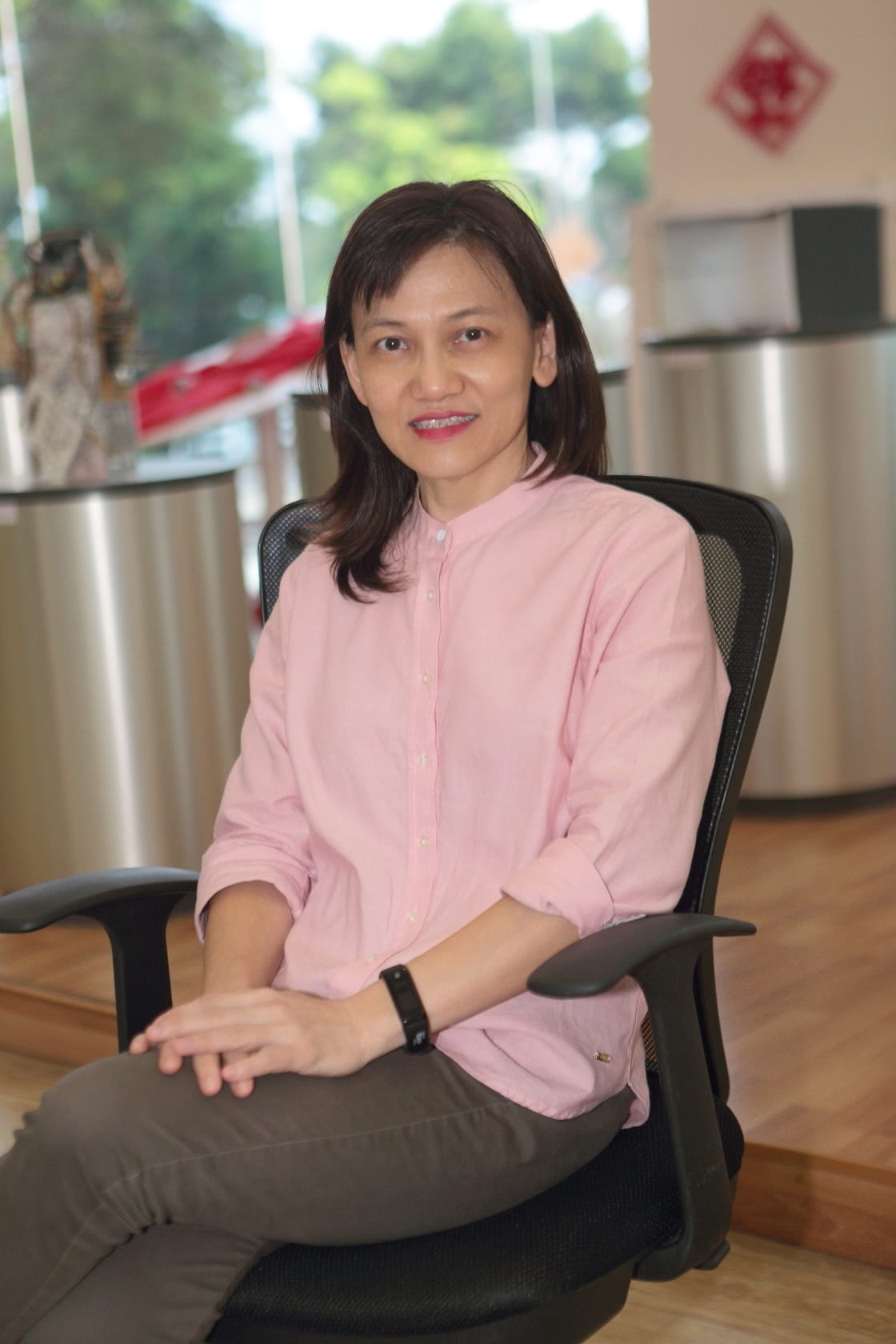 A woman with dark hair in a pink shirt and gray pants sits smiling in a black office chair indoors.