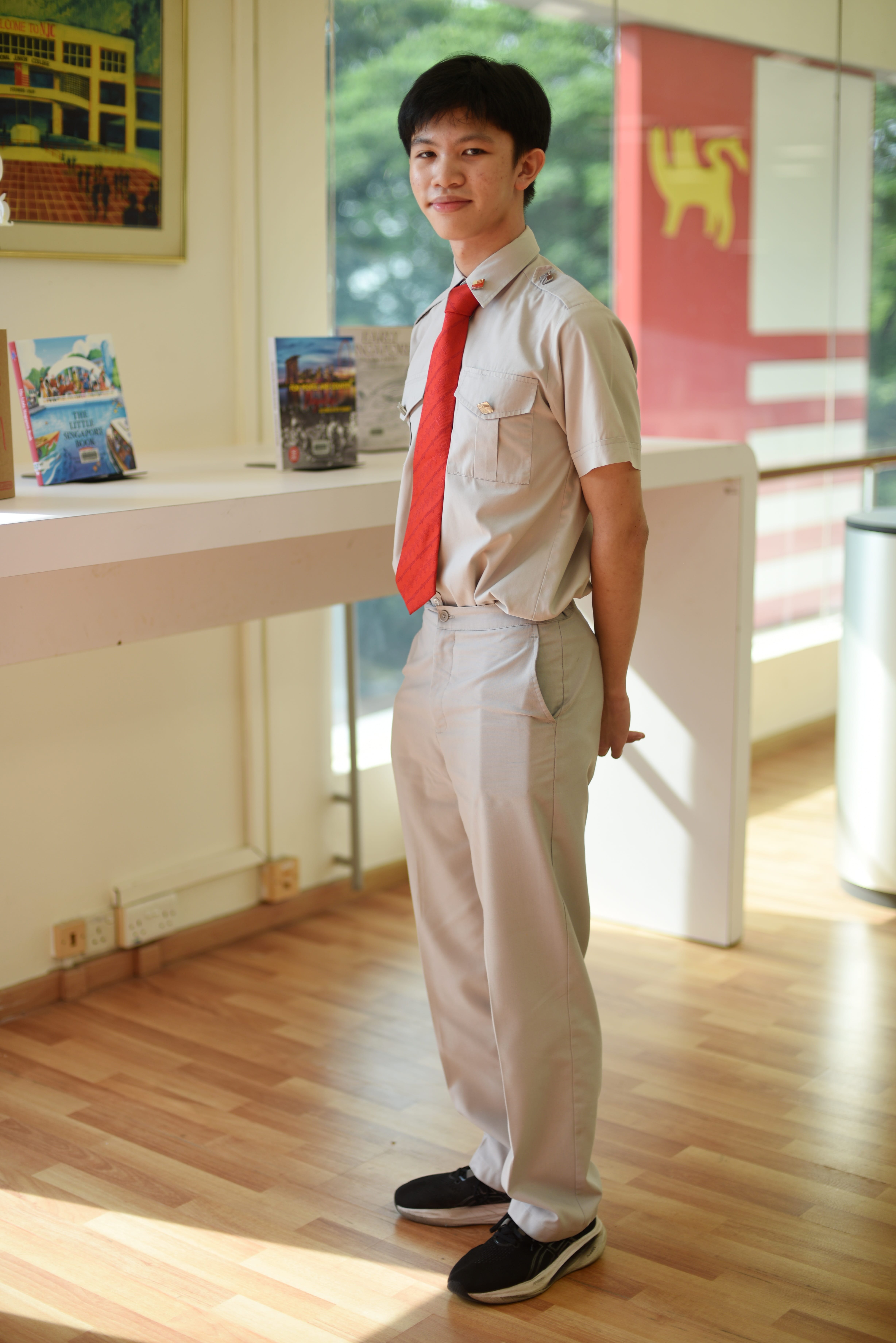 Student in gray uniform with red tie stands indoors by a white table with books, sunlight through windows.