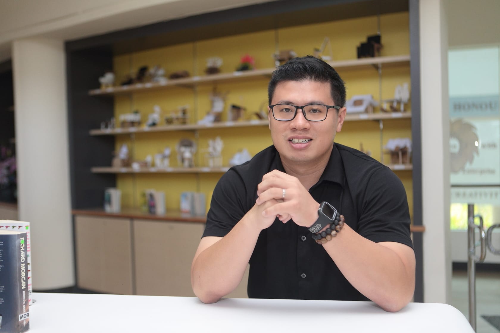 A smiling man in a black shirt and glasses sits at a white table with shelves of small objects behind him.