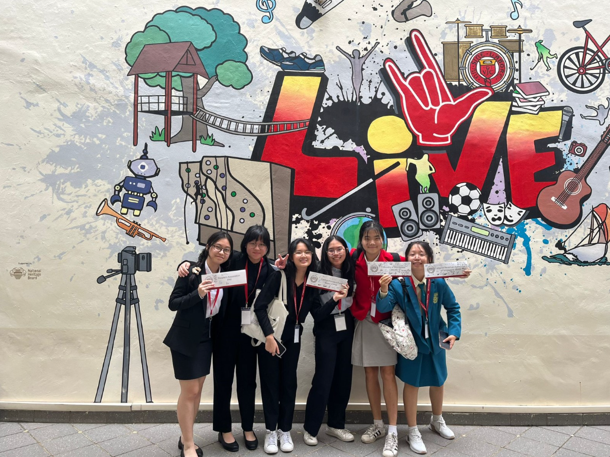 Six young women holding certificates pose smiling in front of a colorful mural with the word LIVE.
