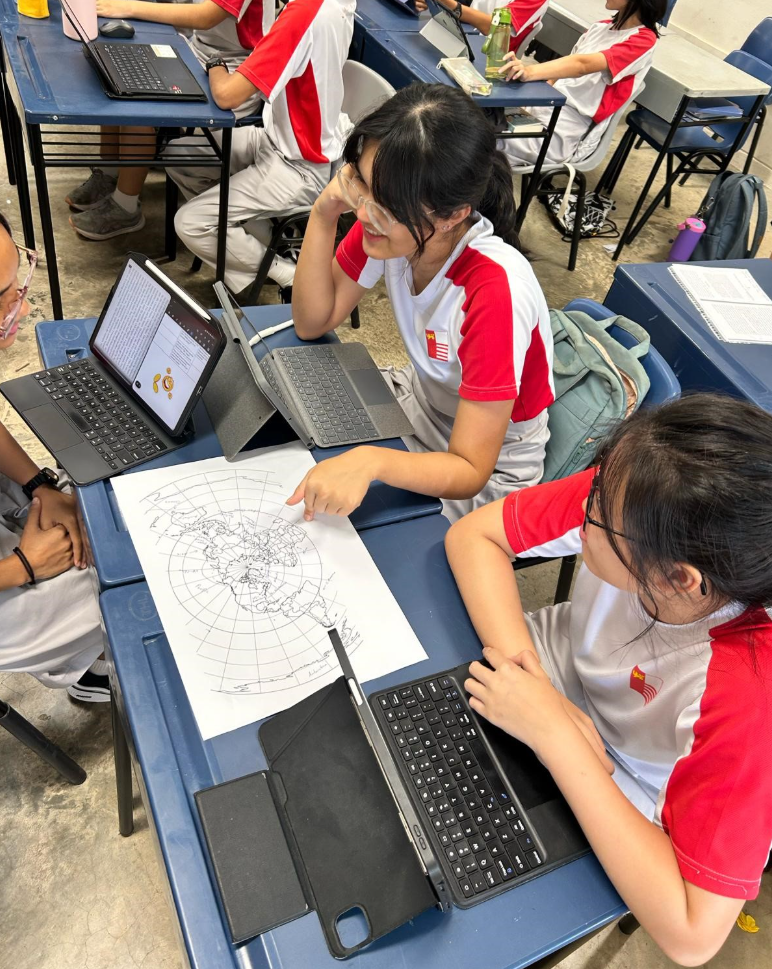 Two students in red and white uniforms examine a printed world map on a desk with tablets nearby in a classroom.