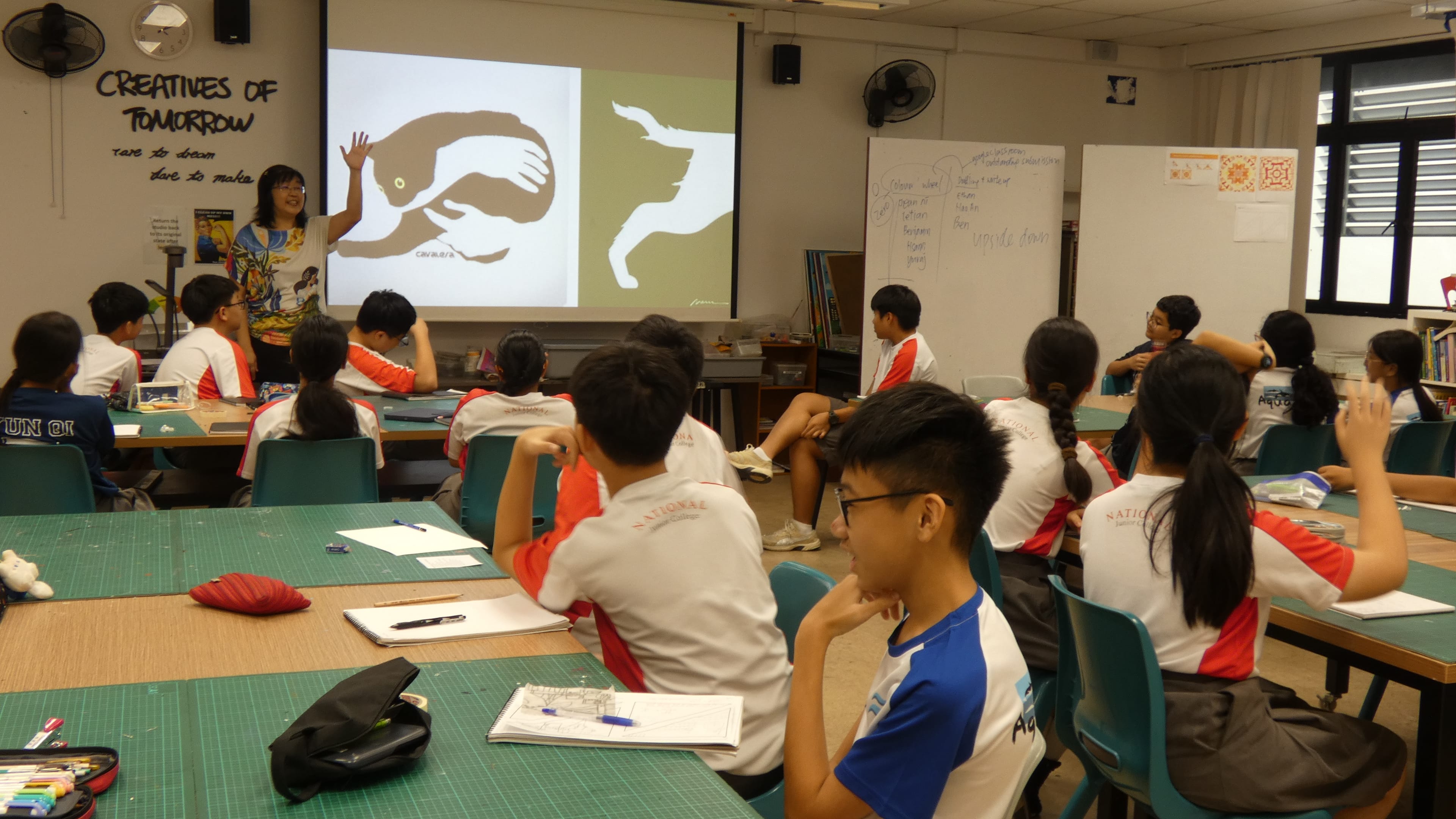 Teacher raises hand by projector screen as students in red-and-white uniforms sit at tables in a classroom.