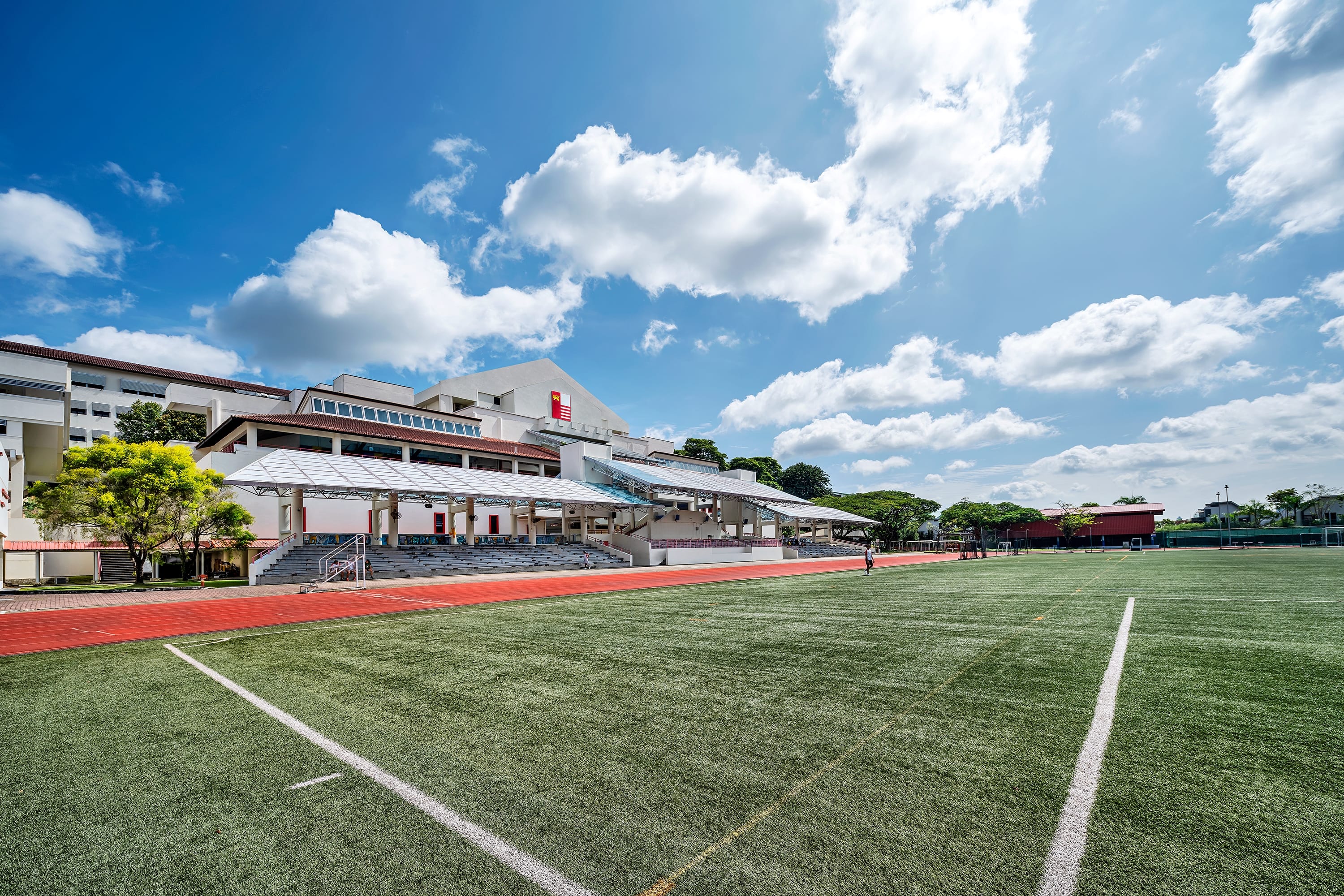 Sports field with white lines and red track beside covered stadium seating under a blue sky with clouds