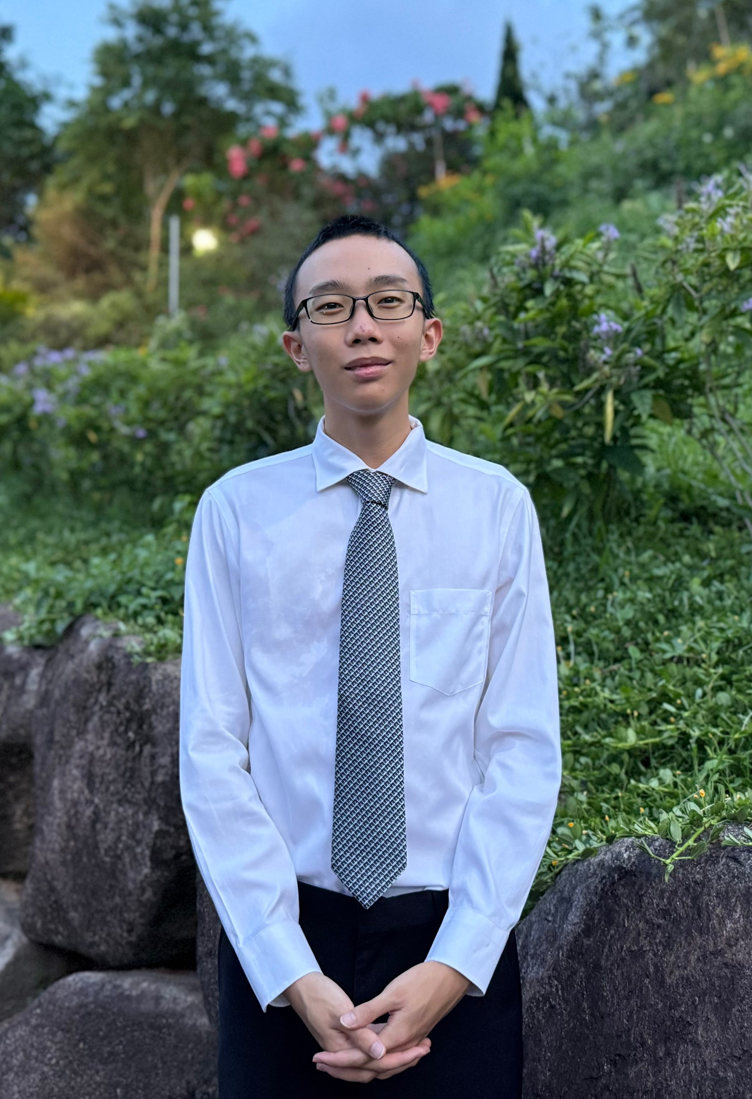 Young man with glasses wearing a white dress shirt and patterned tie, standing outdoors near rocks and green plants.