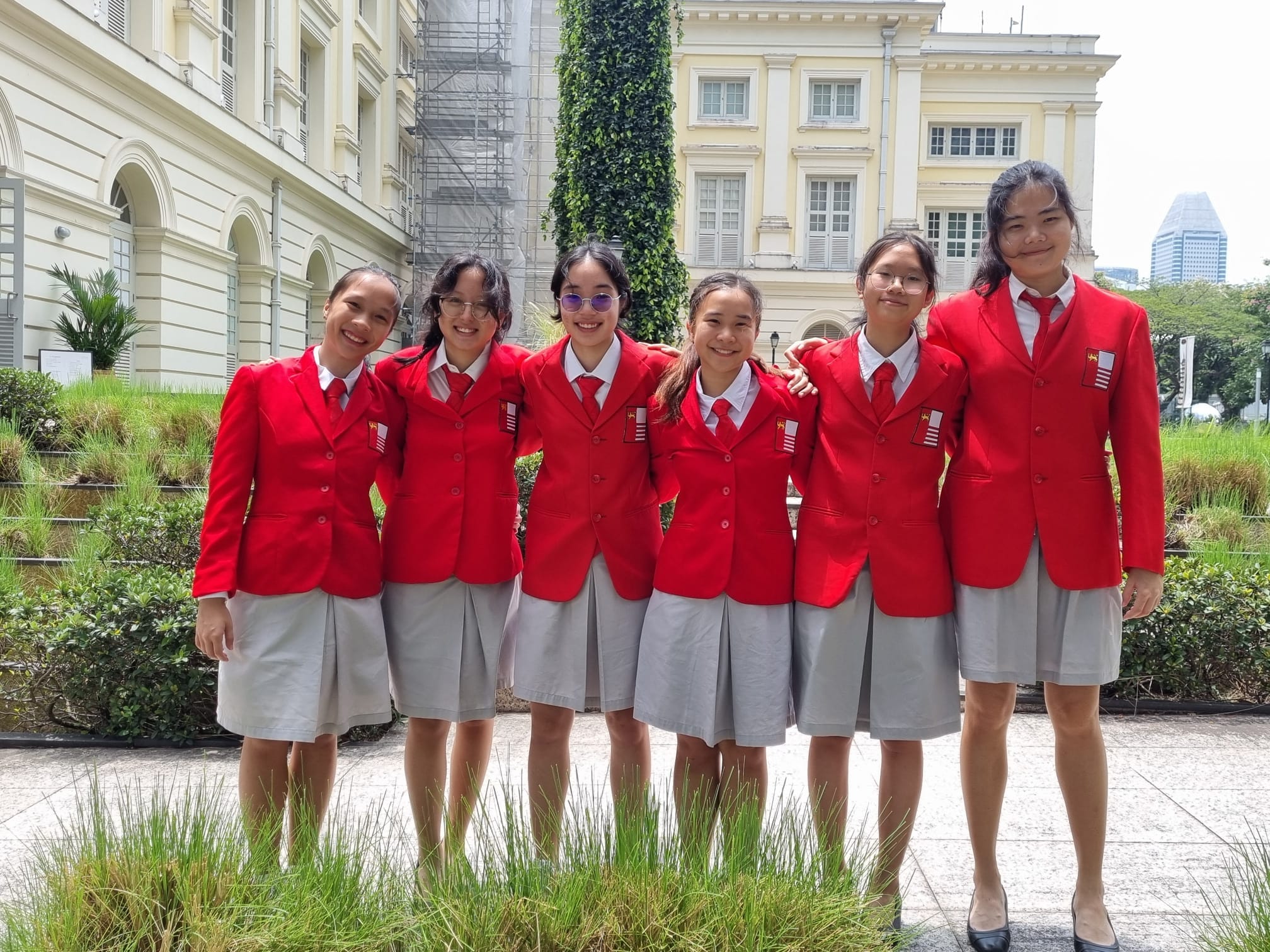 Six girls in red blazers and gray skirts pose smiling outdoors in front of a classical colonial building.