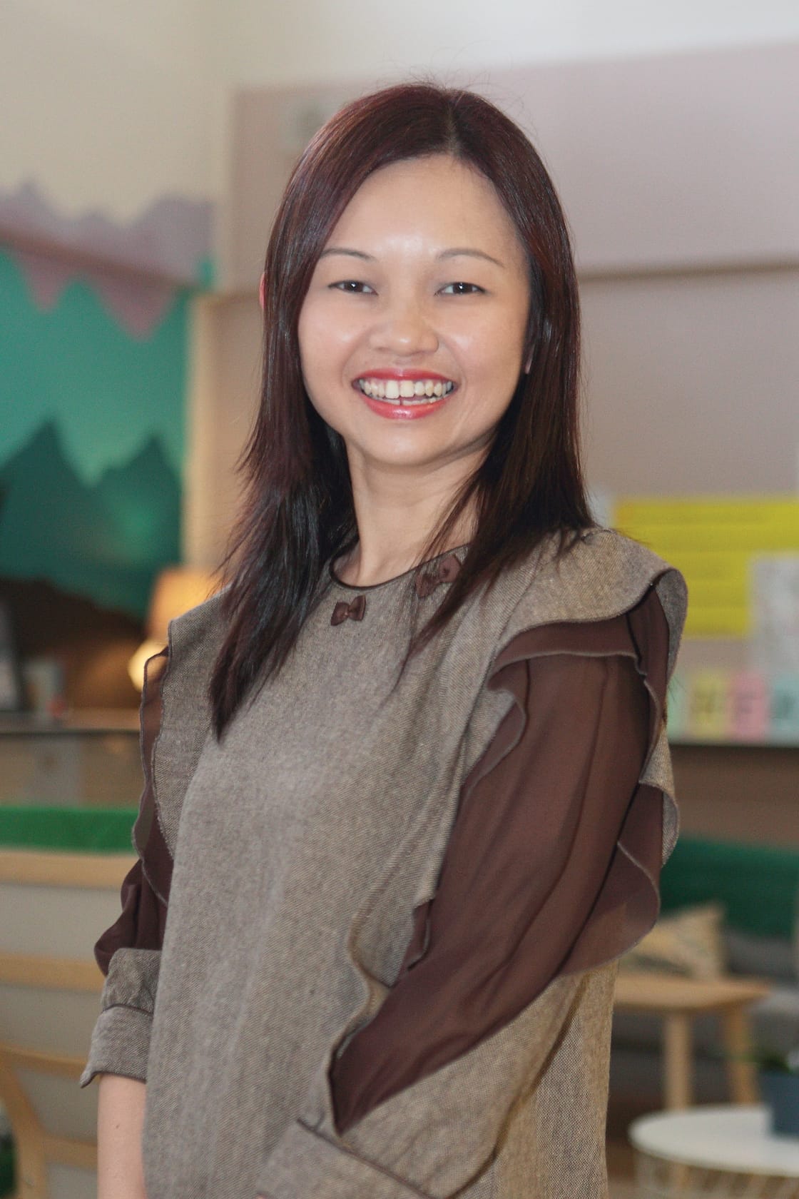 A smiling woman with long dark hair wearing a gray and brown ruffled top, standing in a bright indoor room.