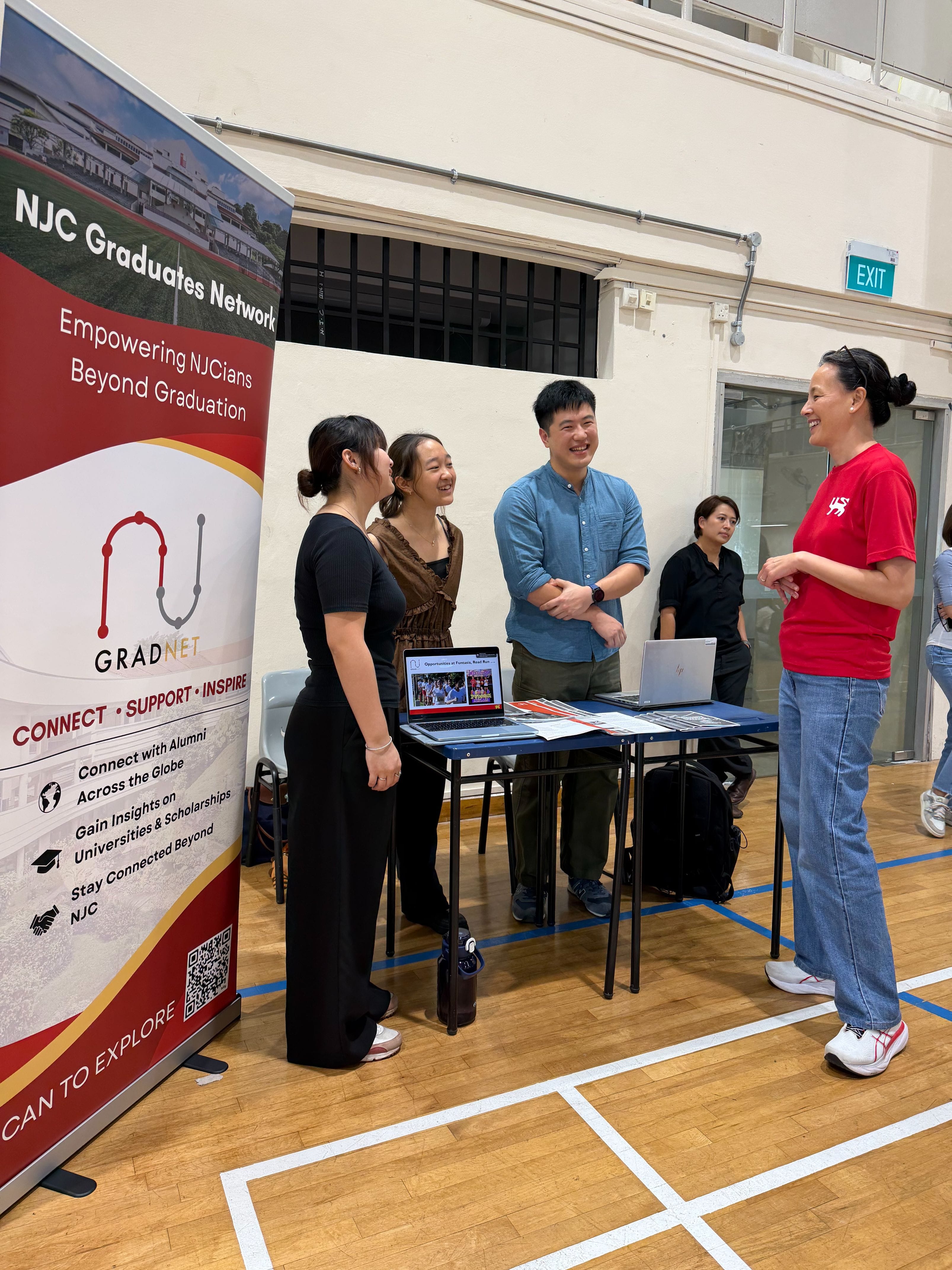 Four people chat at an NJC Graduates Network booth with laptops on a table inside a school hall.