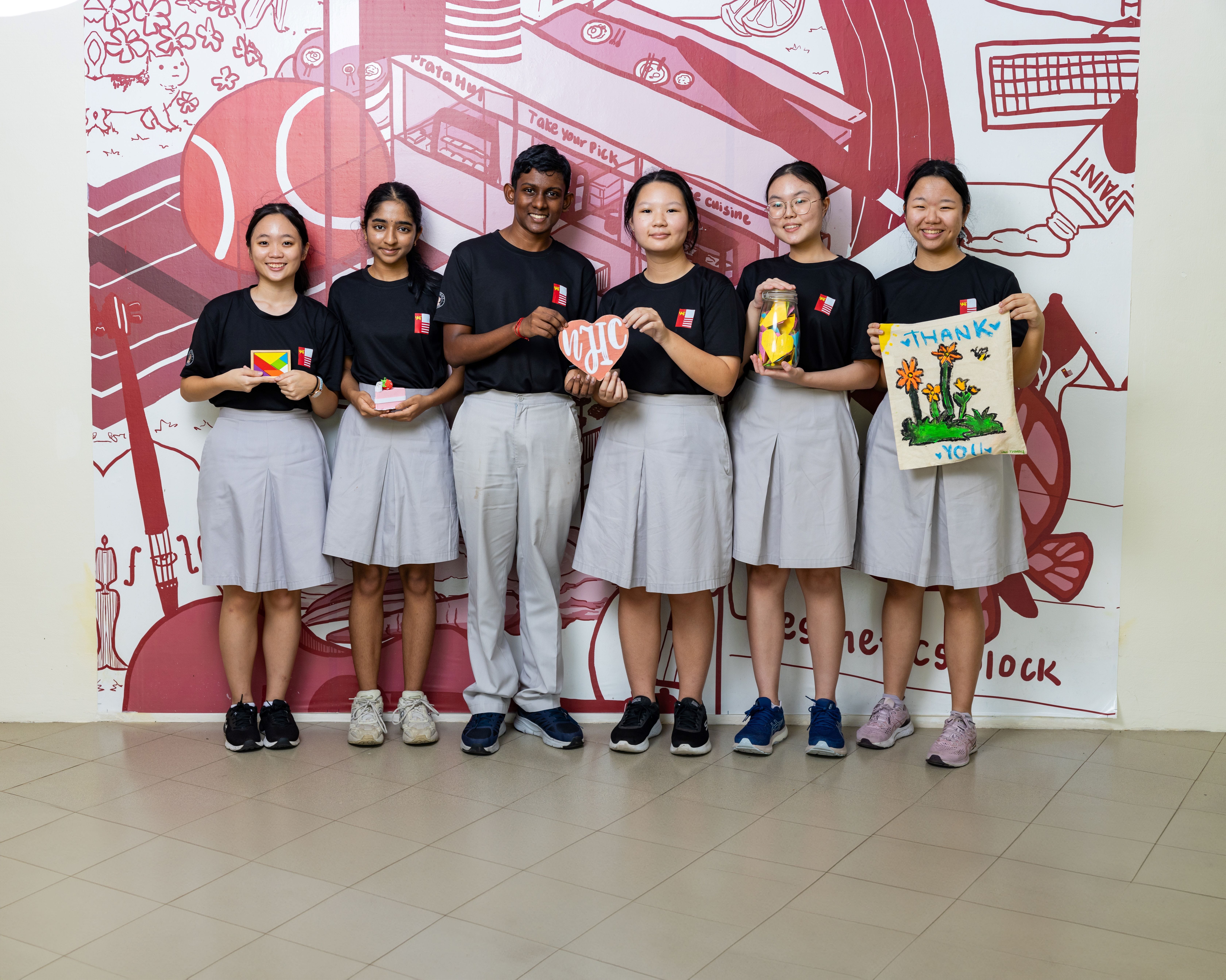Six students in black shirts and grey skirts hold handmade crafts and a thank you poster in front of a red mural.