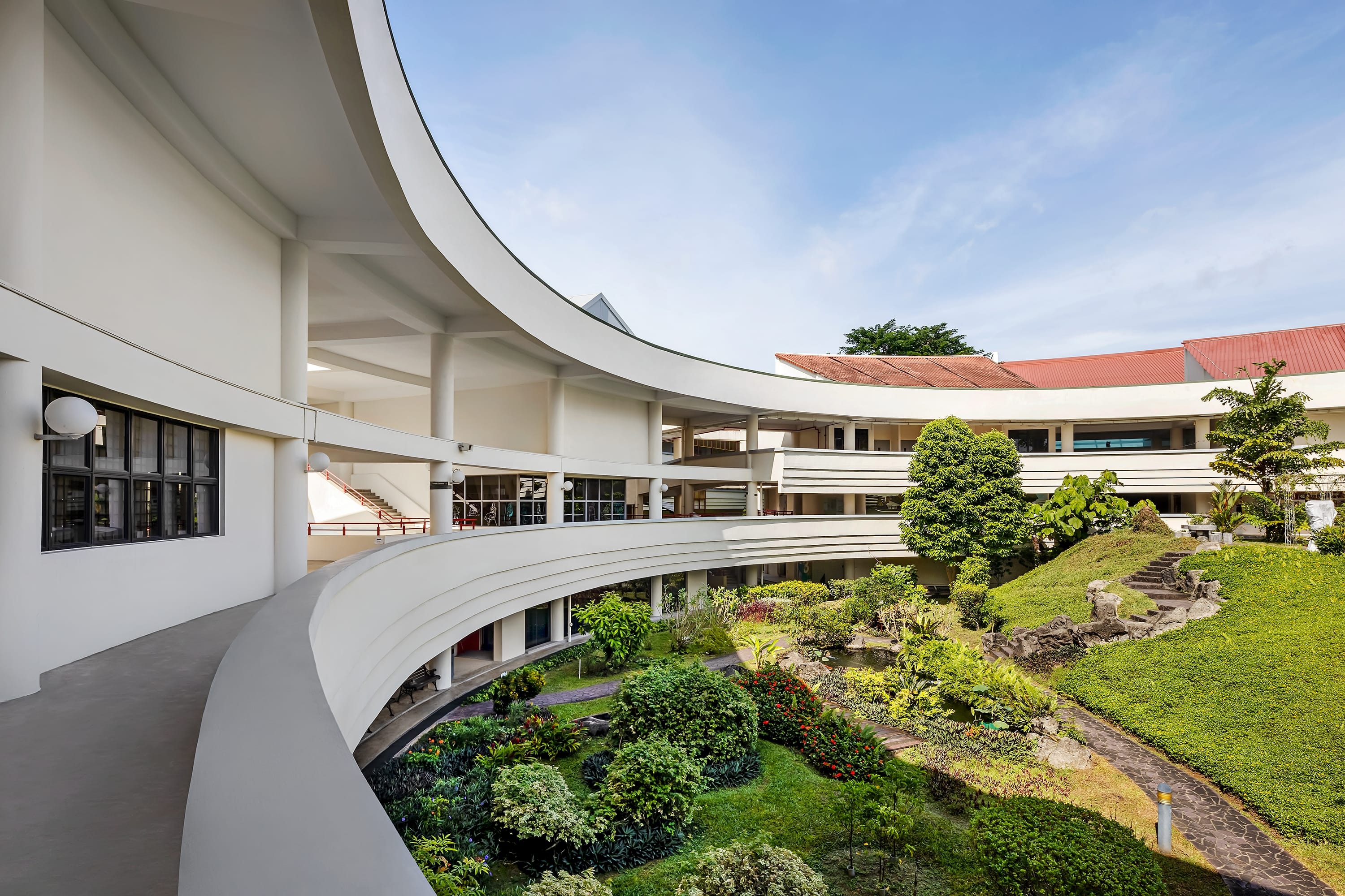 Curved white multi-story building surrounds a courtyard garden with paths, shrubs, and a small rocky hill.
