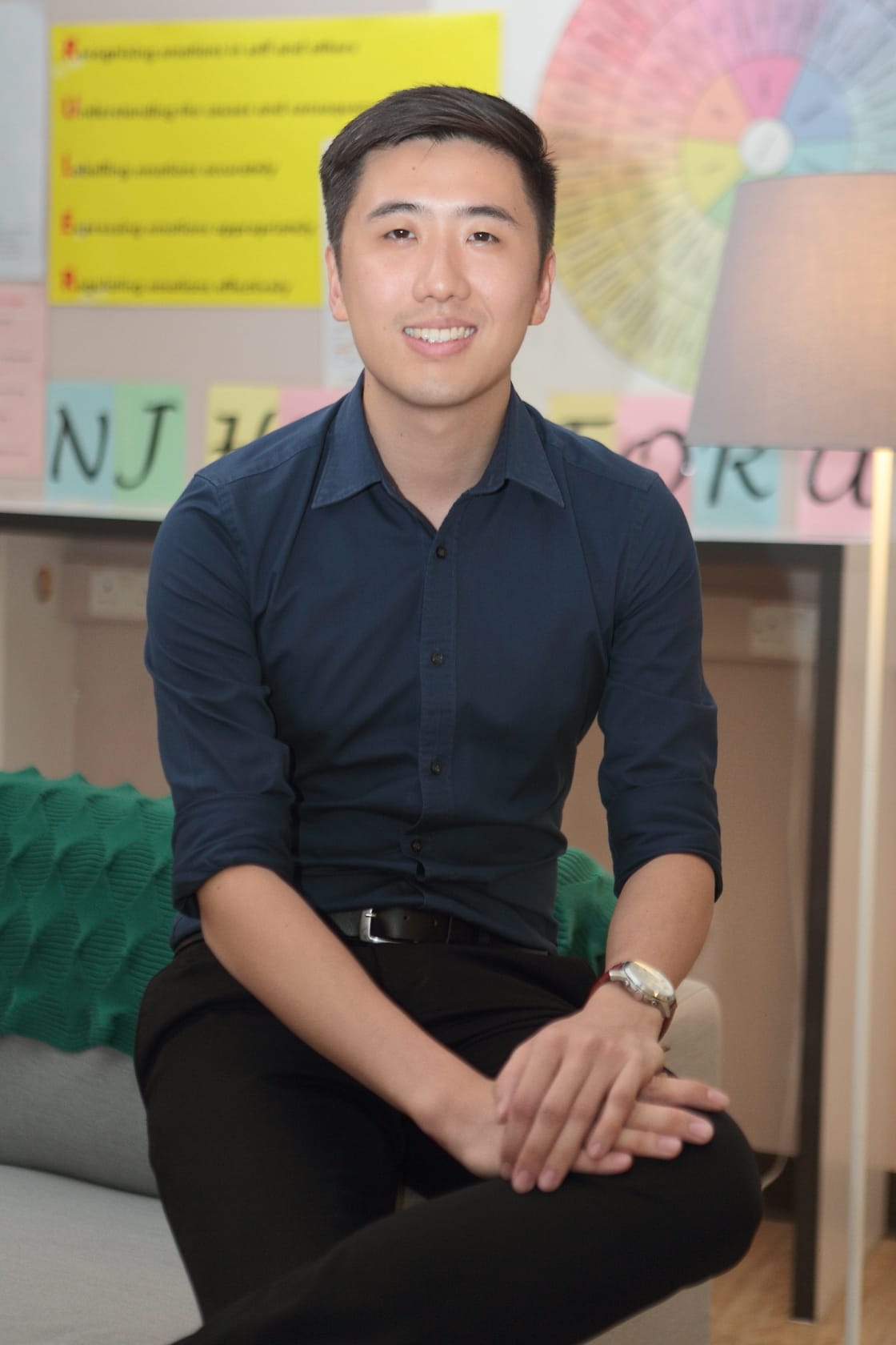 Young man in a navy shirt and black pants sits smiling, with colorful posters and sticky notes on the wall behind him.