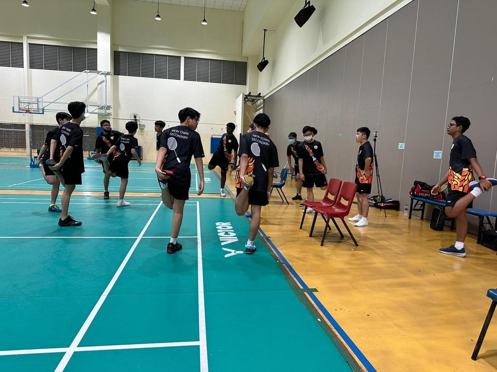 Students stretching to warm up before a badminton match at an external school's indoor sports hall.