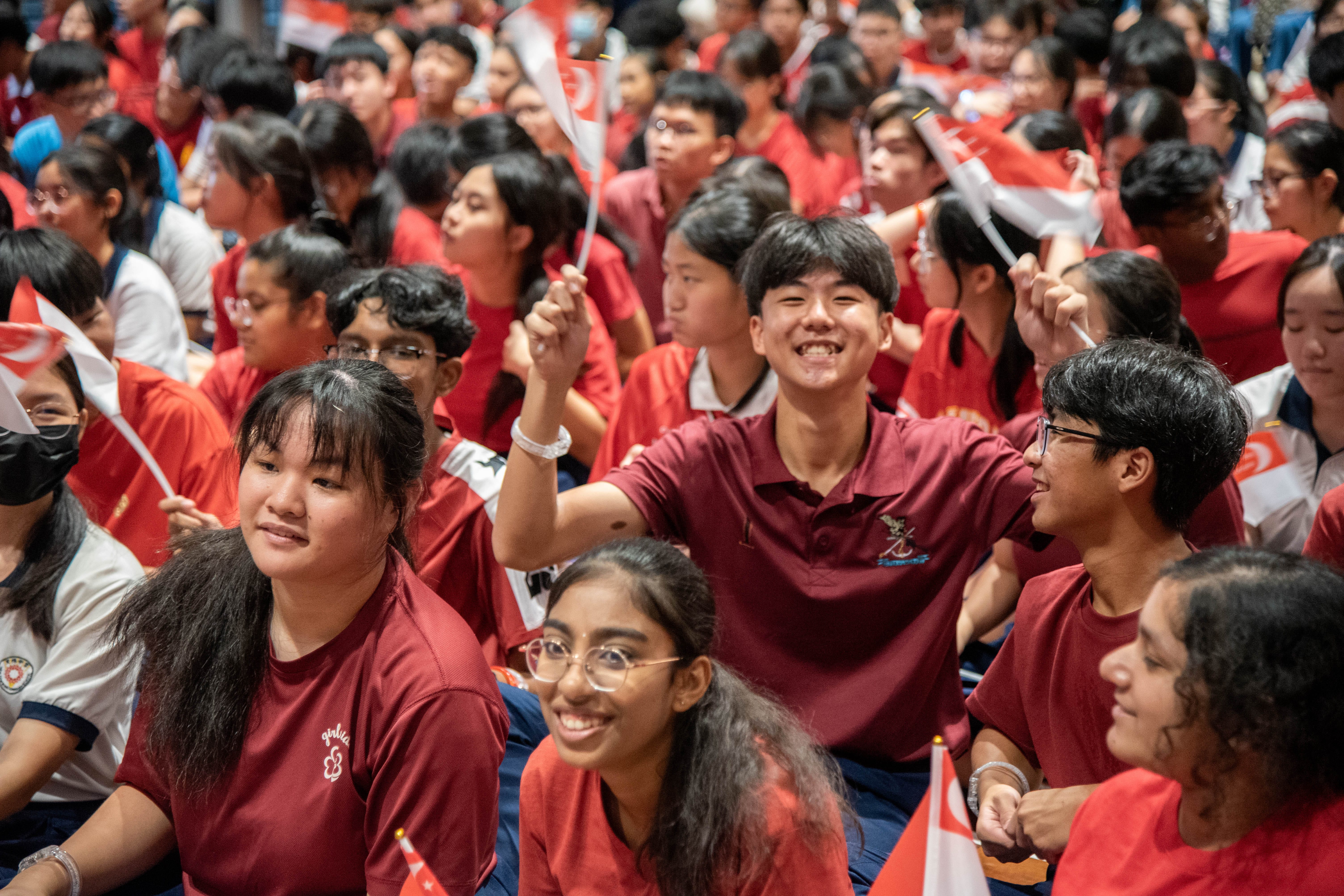 Students happily smiling and excitedly waving their flags as they sit congregated in the school hall to celebrate National Day.