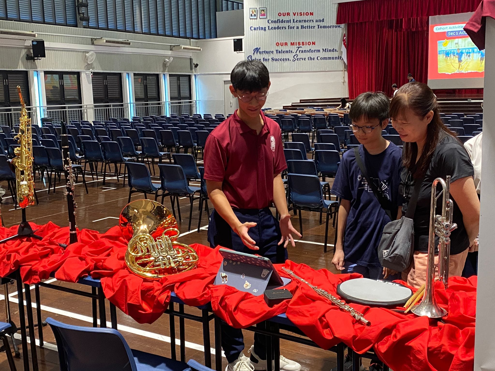Three students stand near a red-draped table displaying a French horn, flute, trumpet, and other instruments in a school hall.