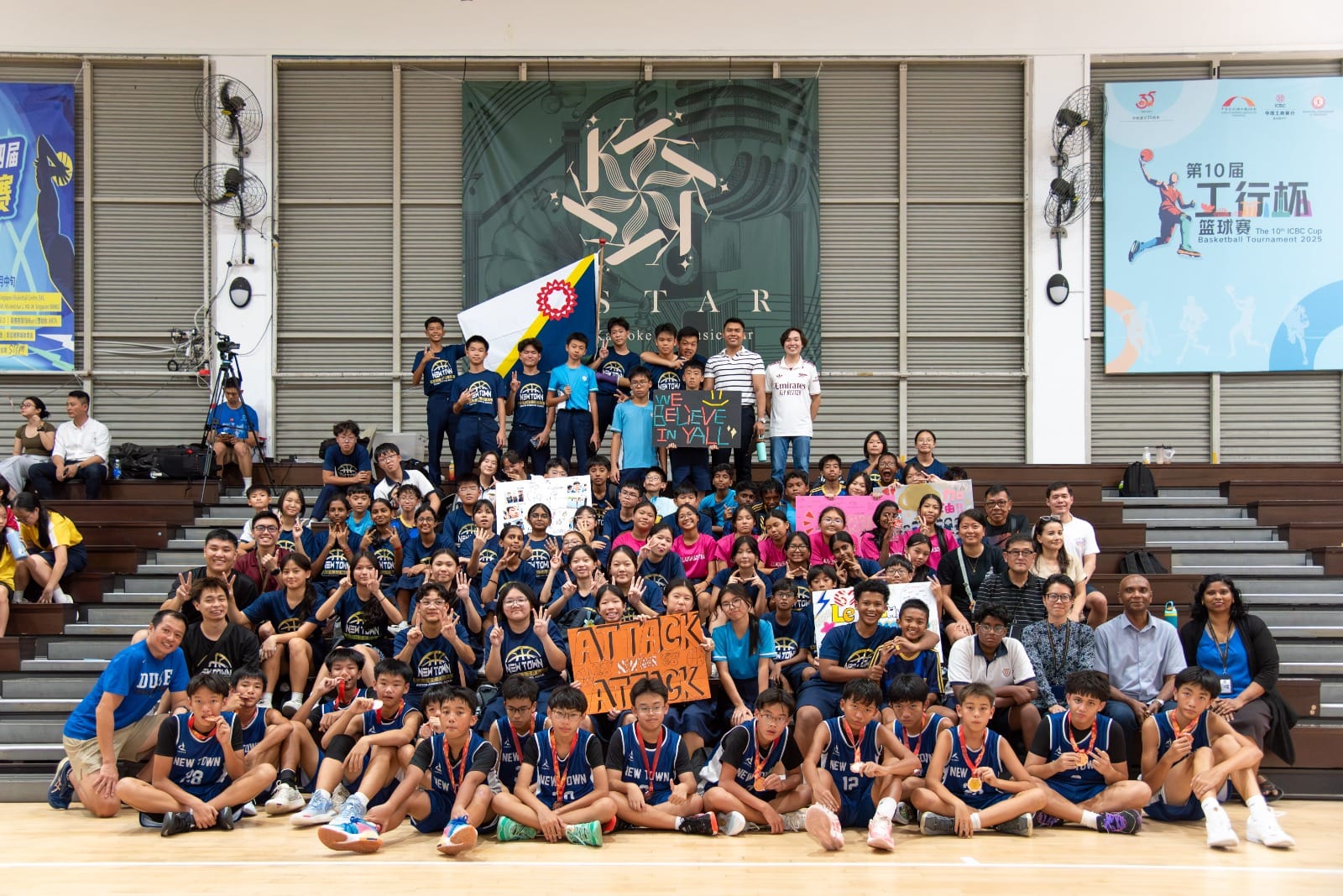Large group of students in New Town basketball jerseys posing on bleachers with signs and a flag in a sports hall.