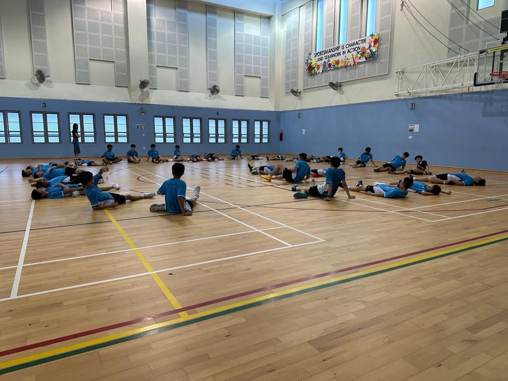 Students stretch and rest on the wooden floor of the indoor sports hall.