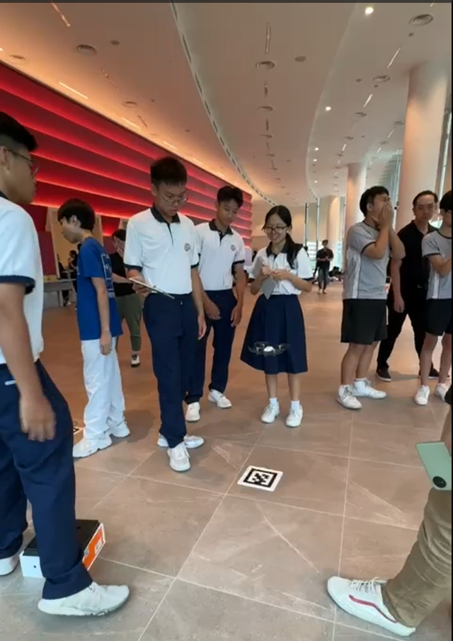 Students in white and navy uniforms stand in a modern indoor hall, with a small drone on the floor nearby.