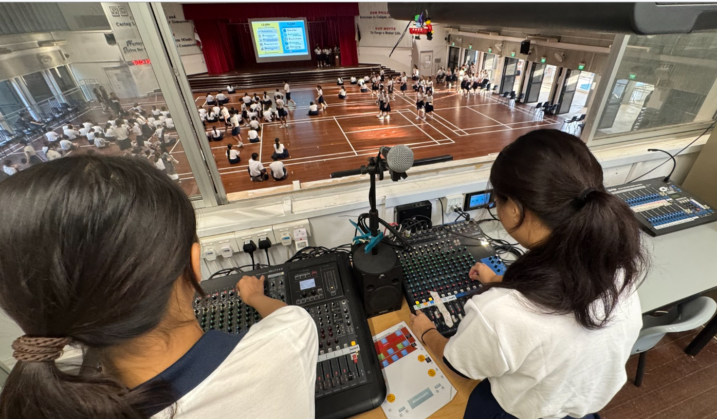 Infocomm club students operate audio mixing consoles in a booth overlooking a school hall filled with uniformed students.