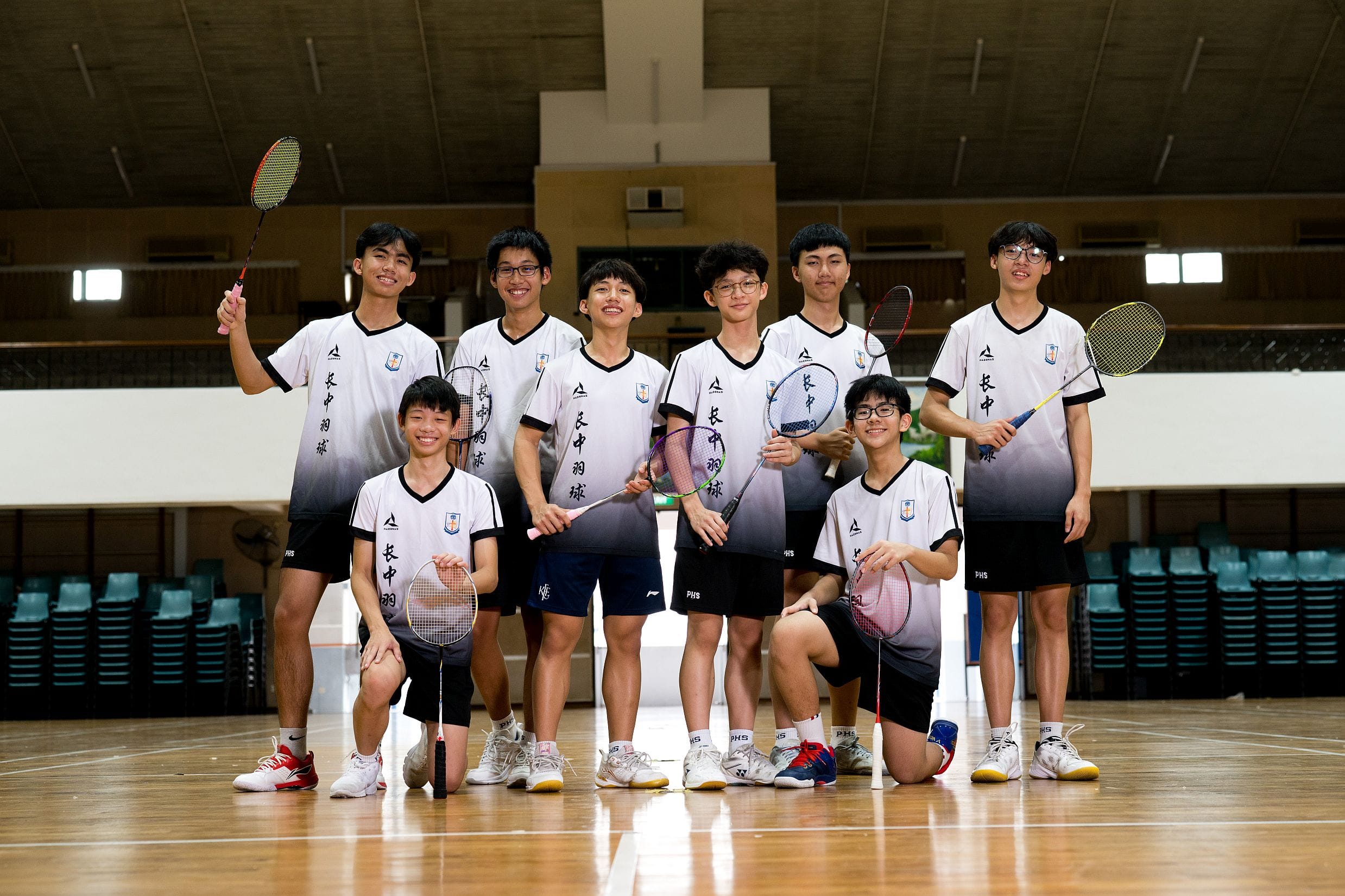 Eight young male badminton players in matching white jerseys pose with rackets inside an indoor sports hall.