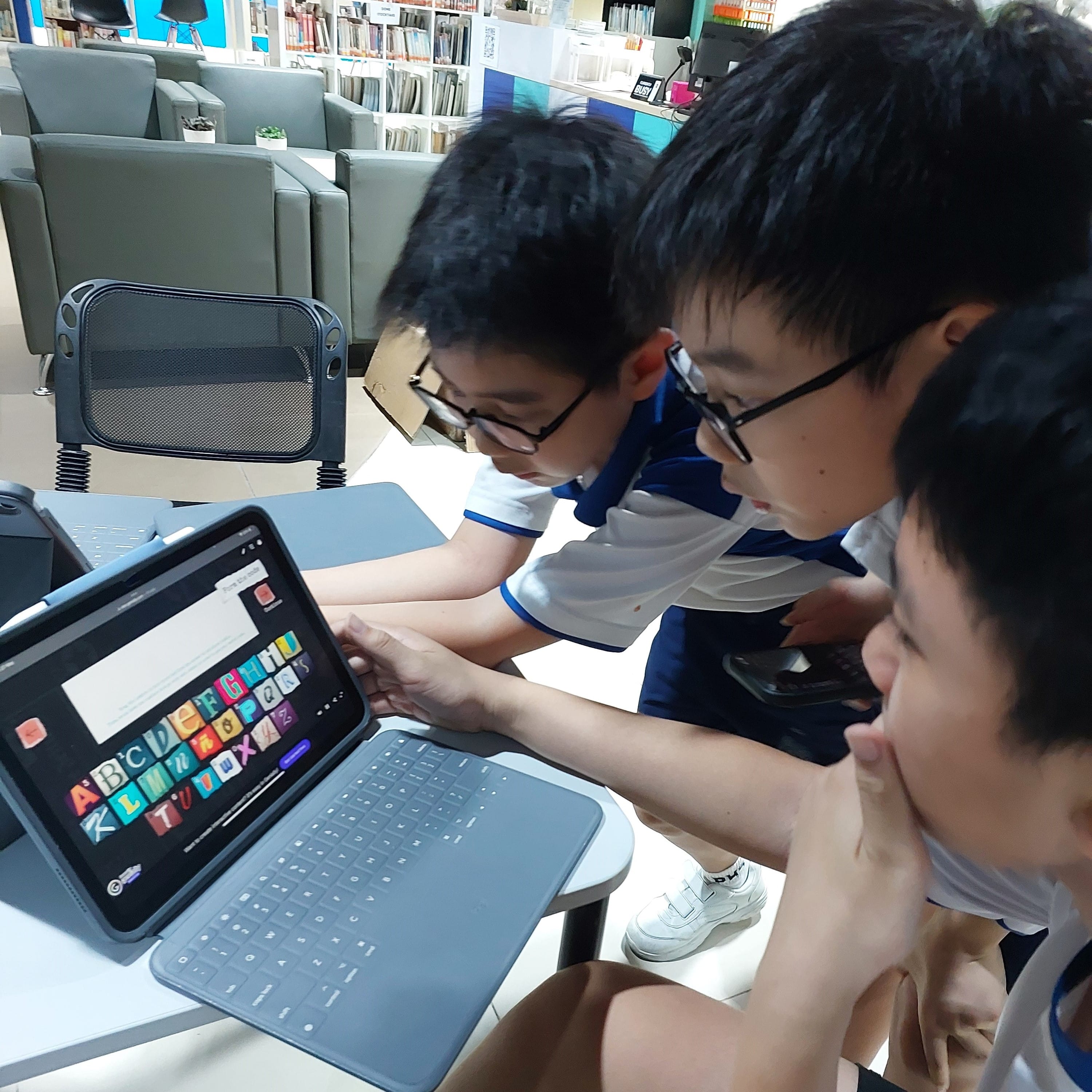 Three boys in school uniforms look at a tablet showing colorful alphabet letters in a library setting.