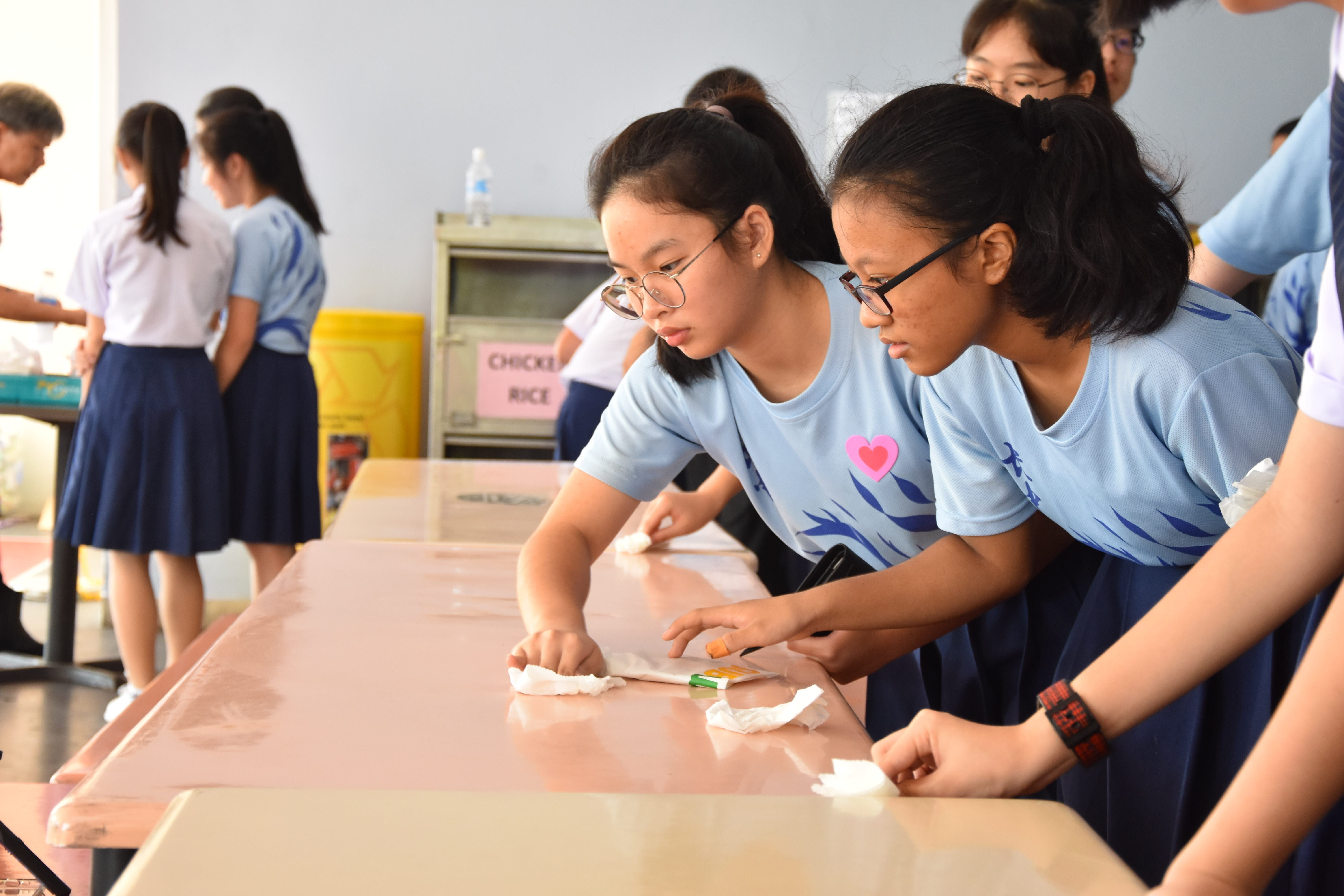 Two girls in blue shirts wearing glasses wipe down a long table in a school cafeteria.