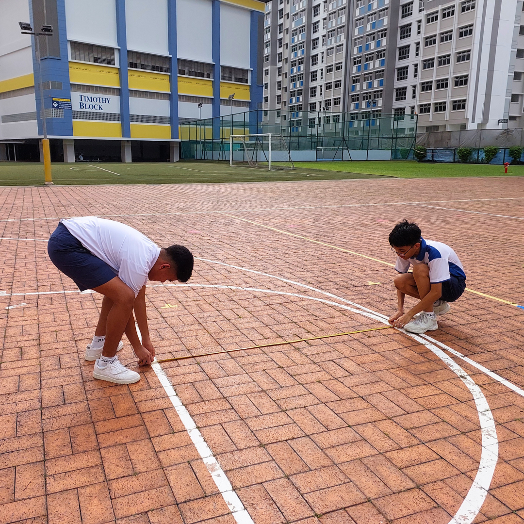 Two boys in white shirts measure a distance on a brick court outside Timothy Block school building.