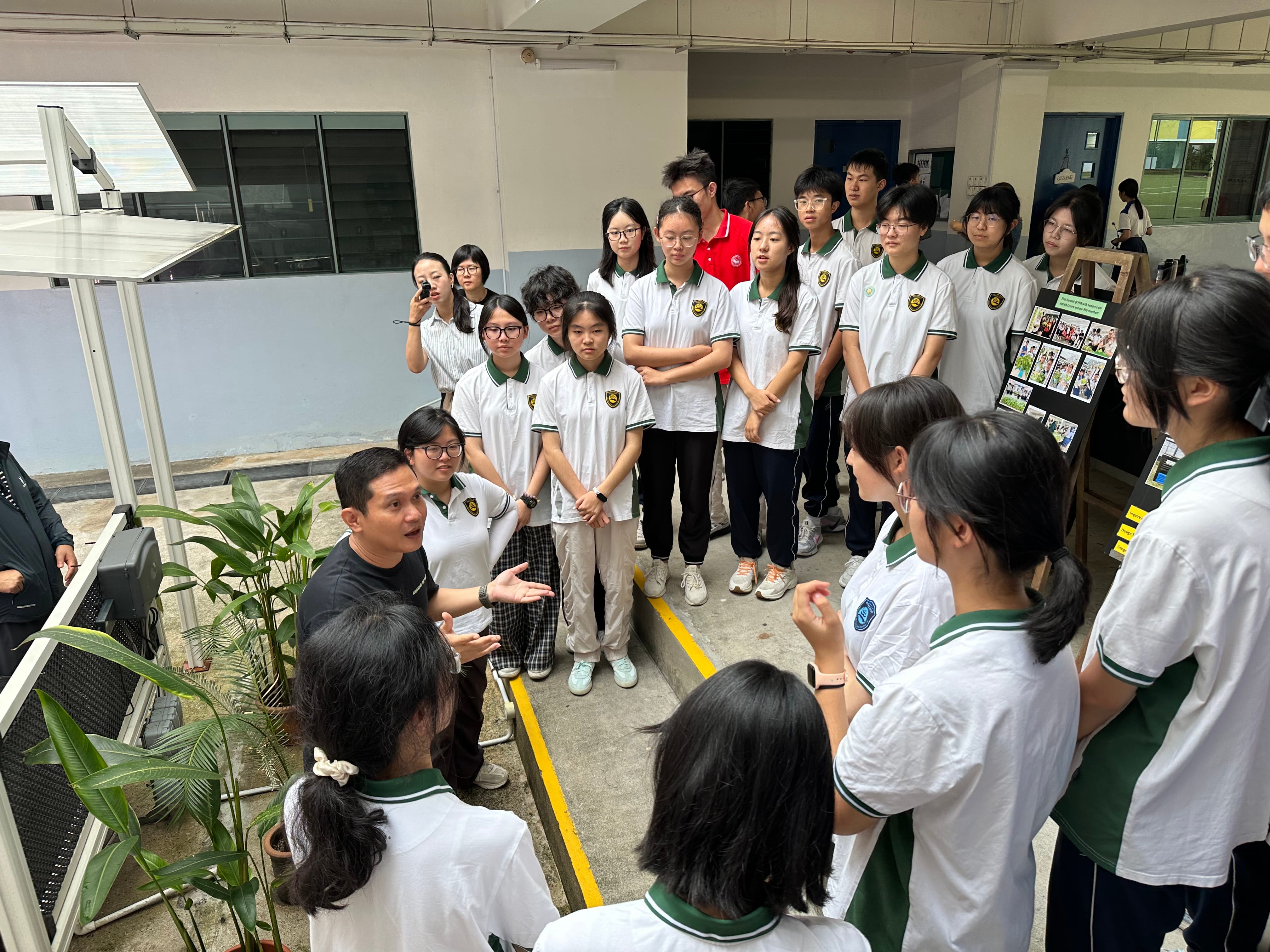 A man in black speaks to a large group of students in white and green uniforms gathered in a school corridor.