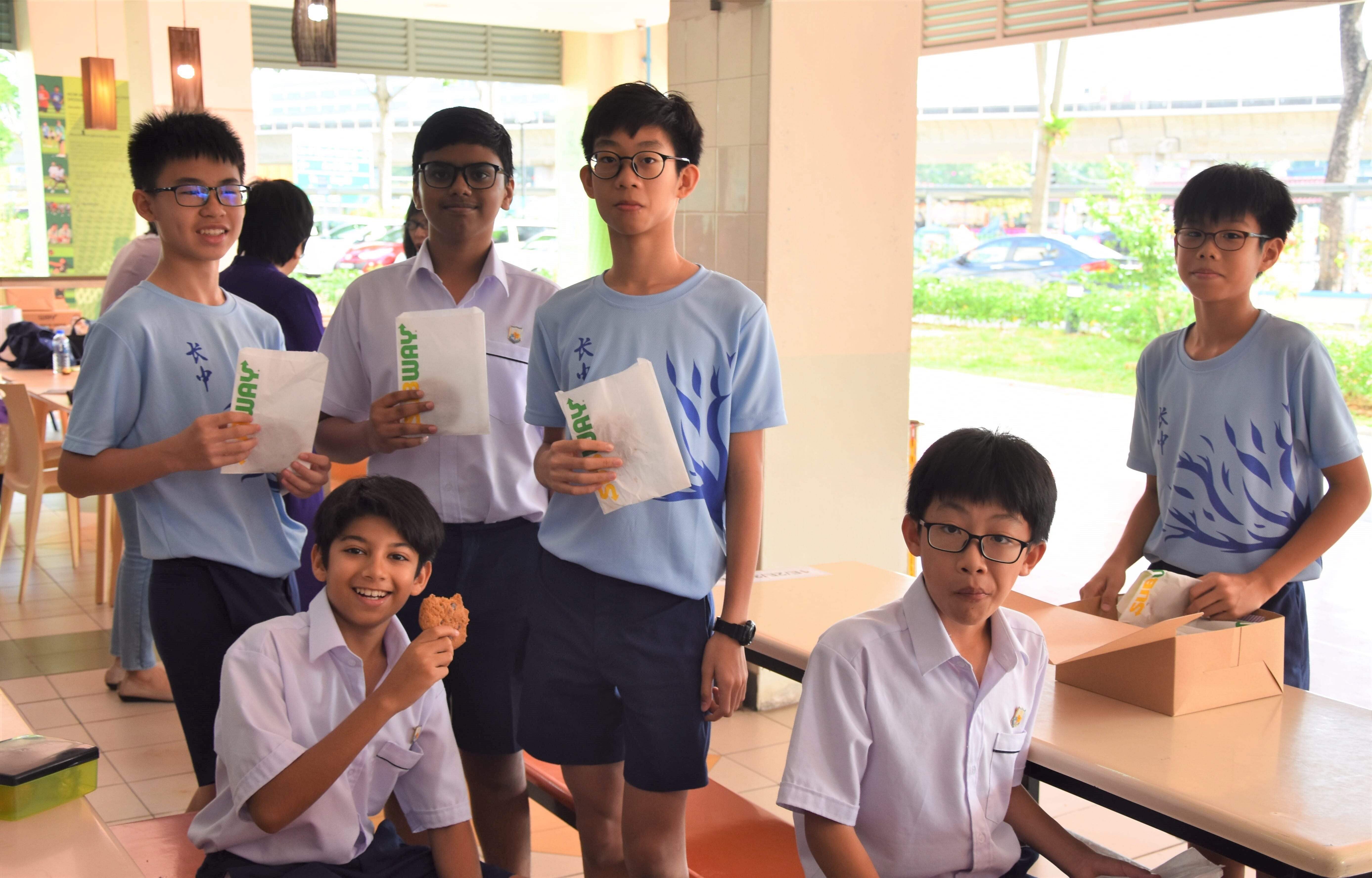 Six school boys in uniforms holding Subway sandwiches and smiling in a school canteen.
