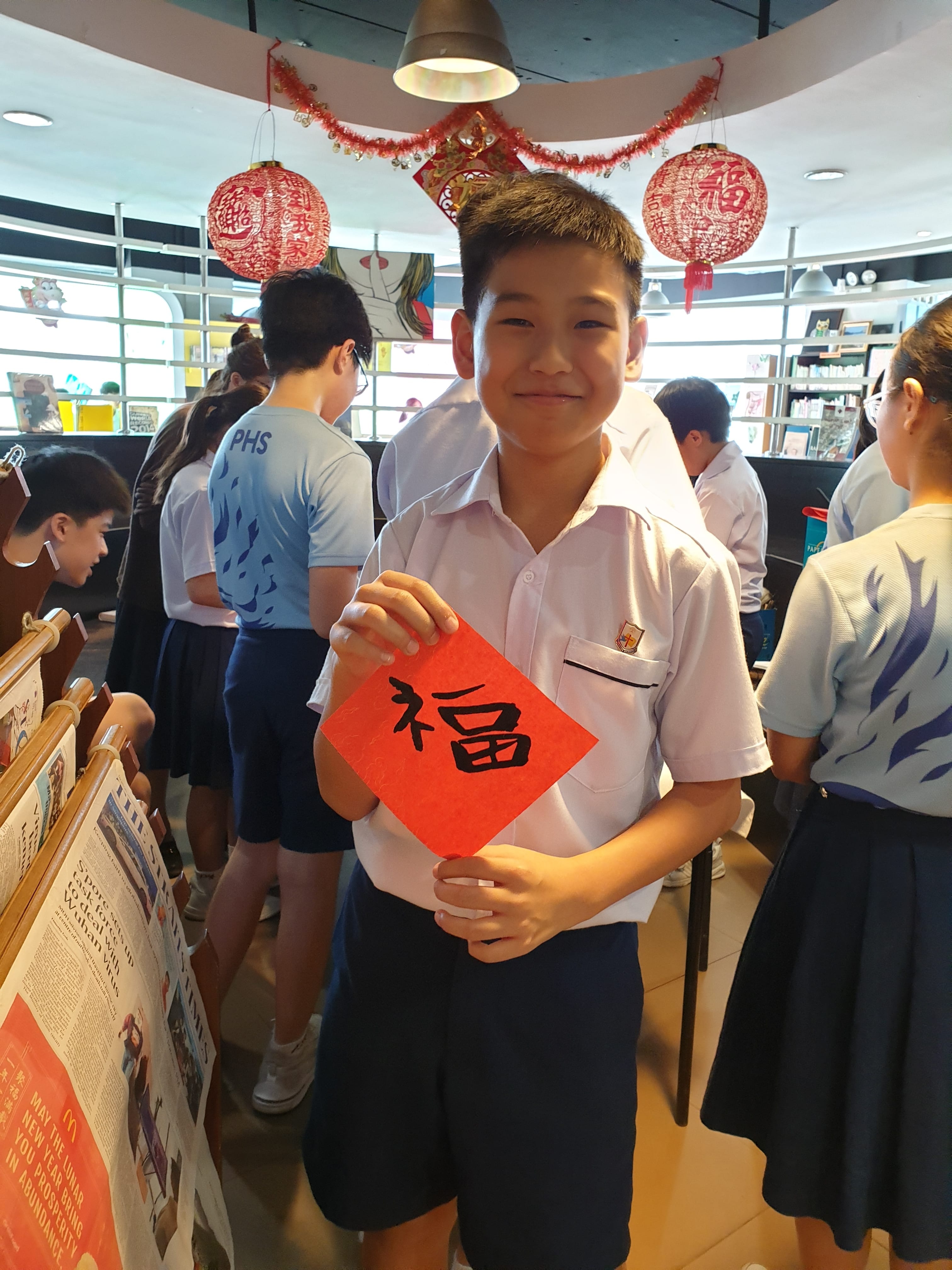 A smiling boy in school uniform holds a red card with the Chinese character Fu, with red lanterns hanging above.