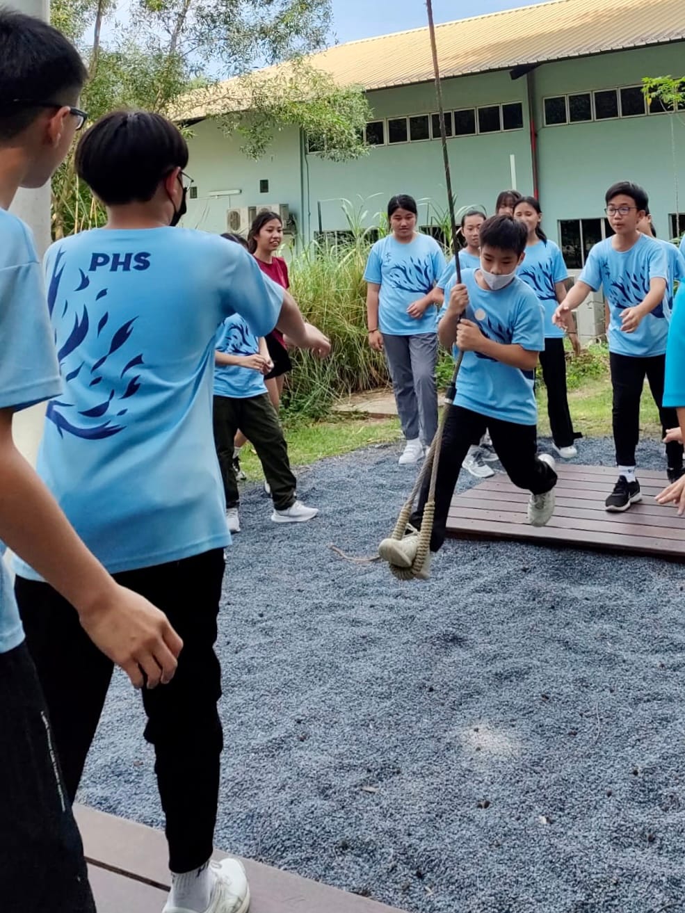 Students in light blue PHS shirts do an outdoor team activity, with one student gripping a rope pole over gravel.