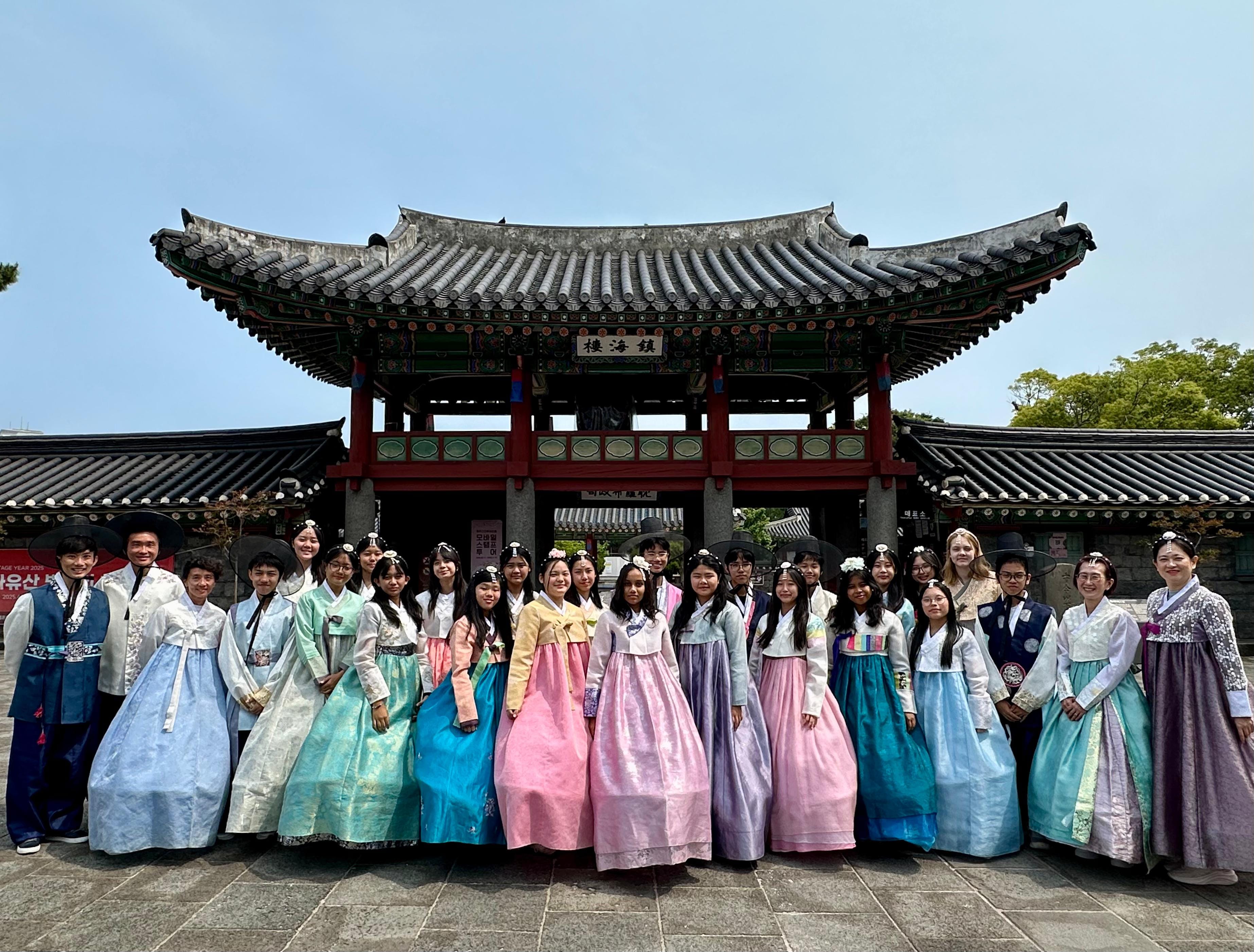 A large group wearing colorful Korean hanbok stands in front of a traditional Korean gate with curved tile roof.