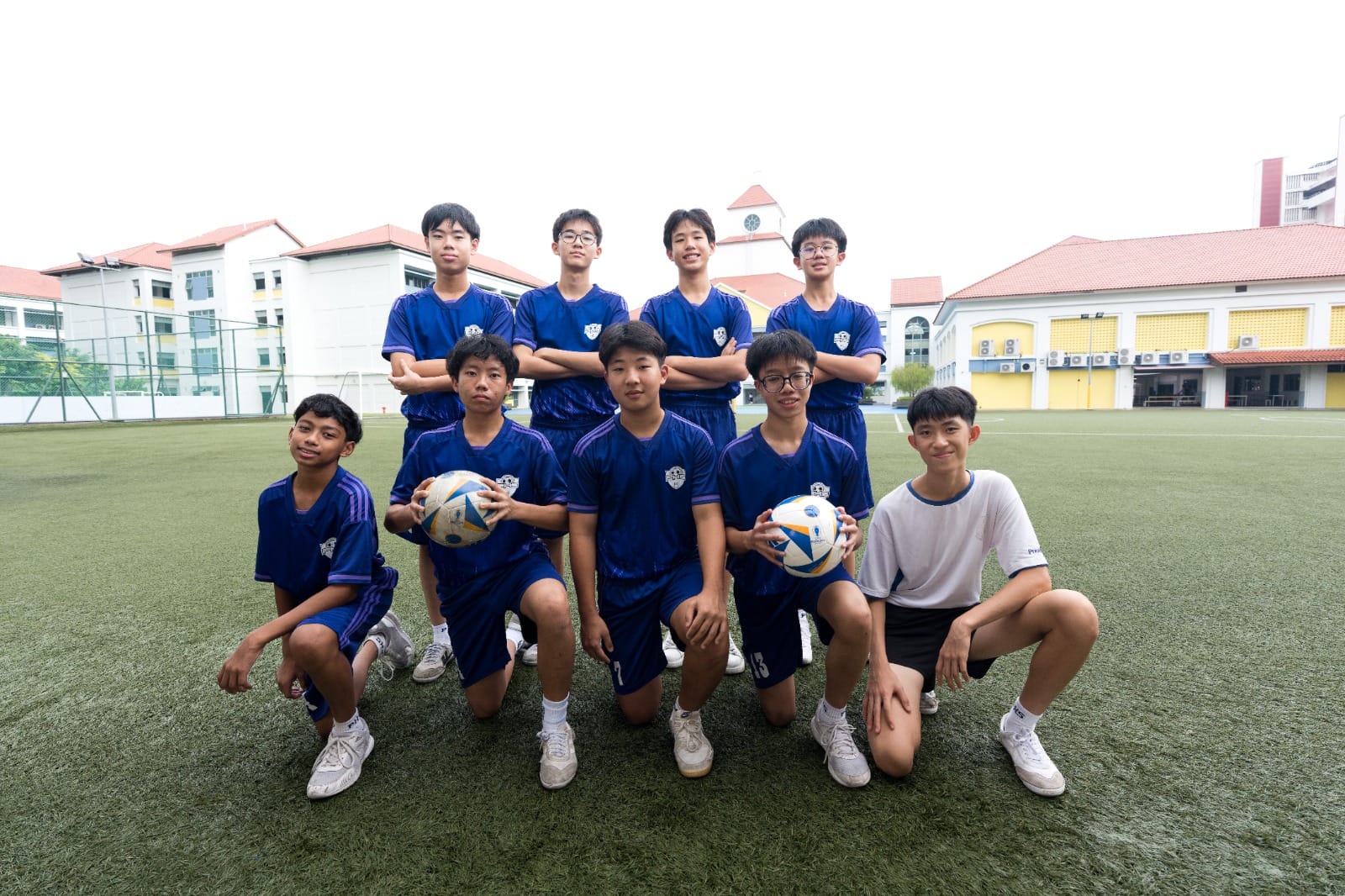 Eight young boys in black football kits pose on a green pitch with a soccer ball, tall apartment buildings in the background.