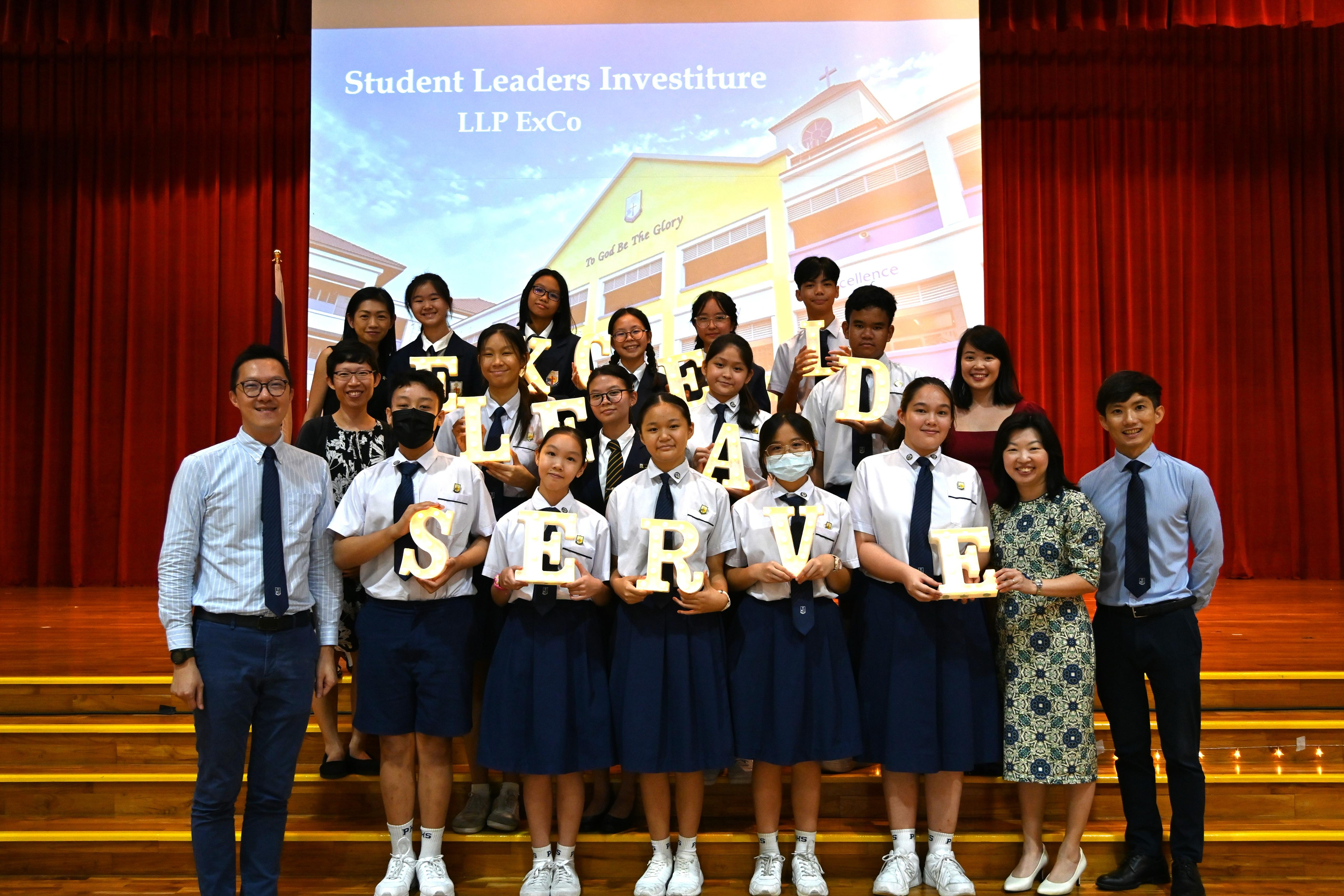 Students and teachers pose on stage at a Student Leaders Investiture, holding lit letters spelling SERVE.