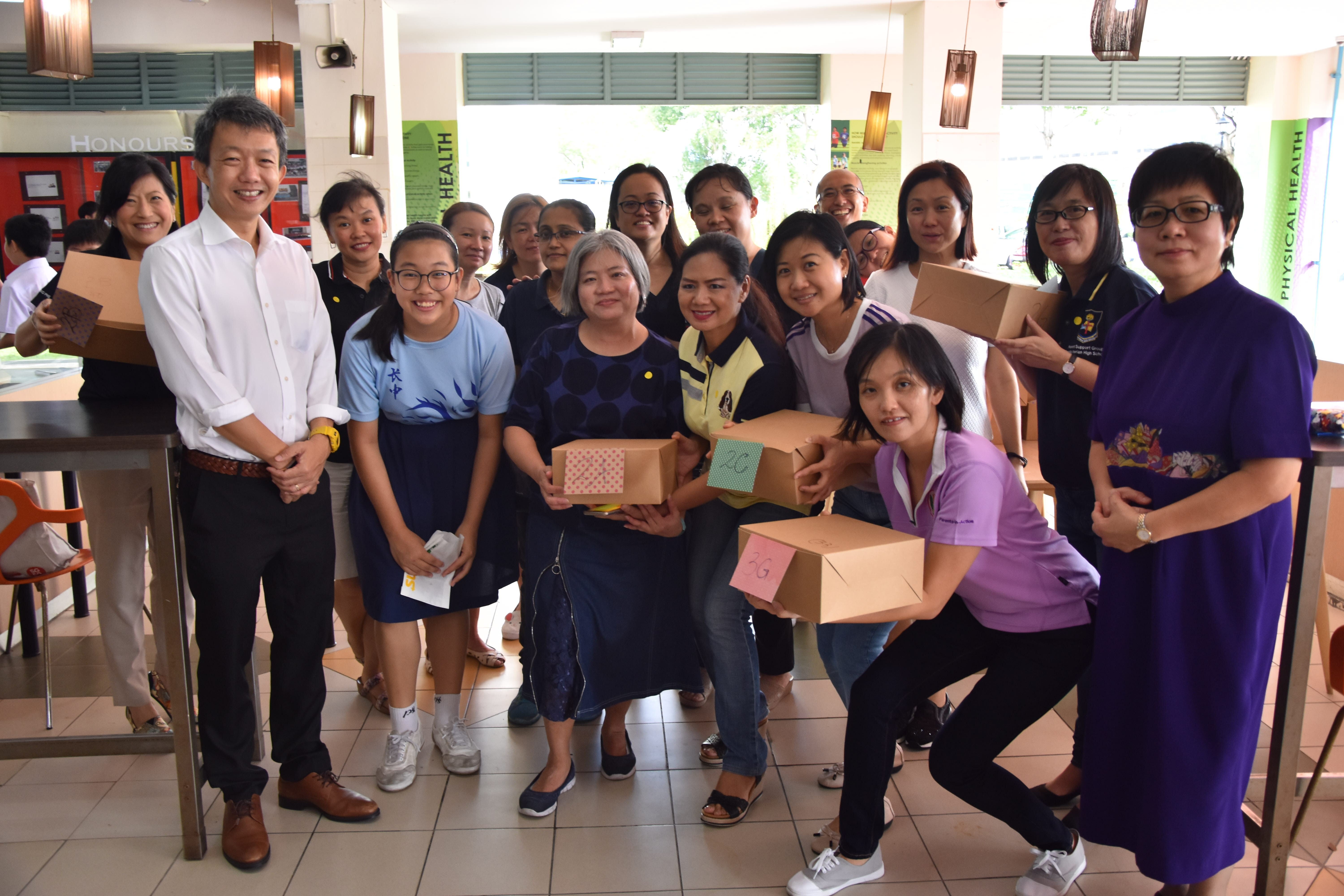 A large group of smiling people pose together indoors, several holding cardboard boxes labeled with letters.