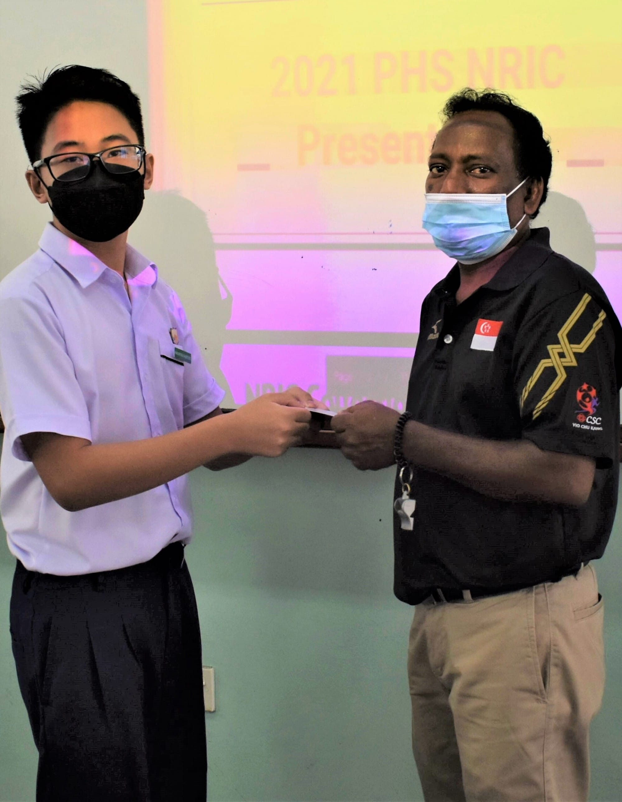 Two masked individuals exchange a document at a 2021 PHS NRIC presentation event indoors.