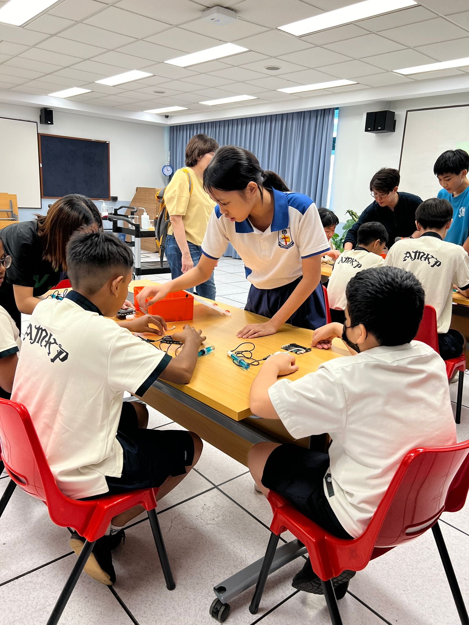 Students in uniforms work on electronics at a classroom table while a girl in a blue skirt leans over to help.