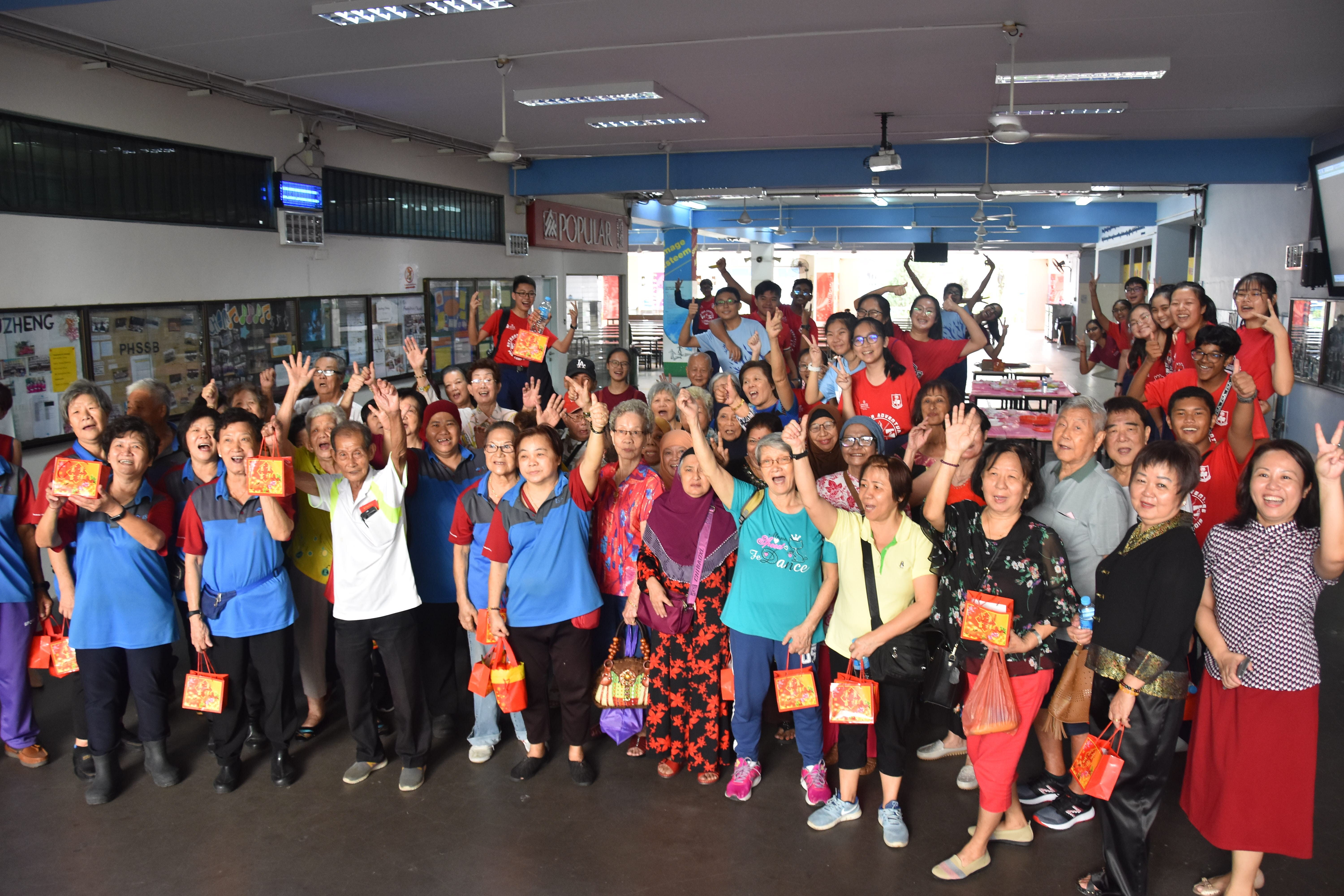 Large group of people of mixed ages smiling and waving indoors, many holding red gift bags.