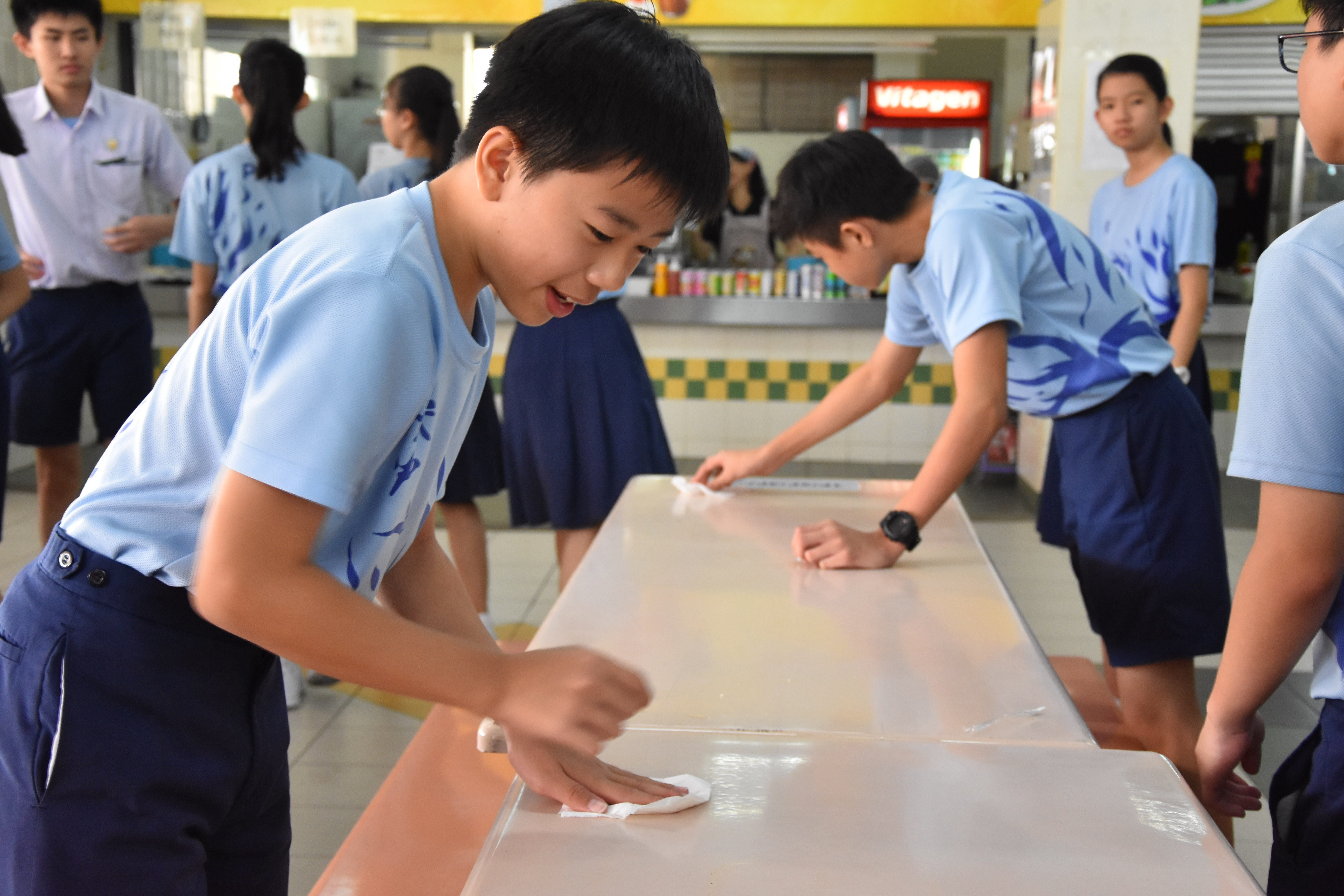 Students in light blue uniforms wiping down a long table in a school canteen.