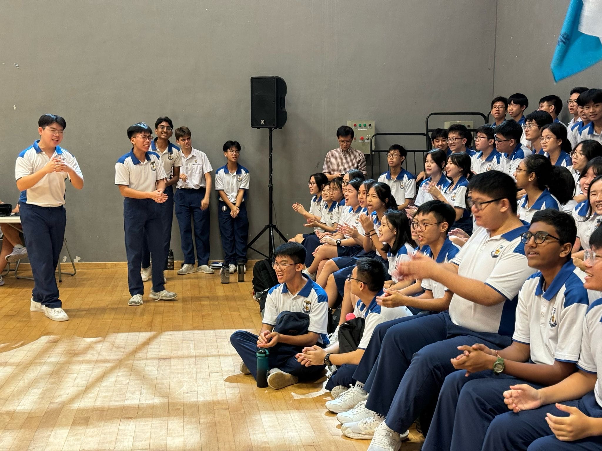 Students in white and blue uniforms gather in a school hall, clapping as two students stand before the group.
