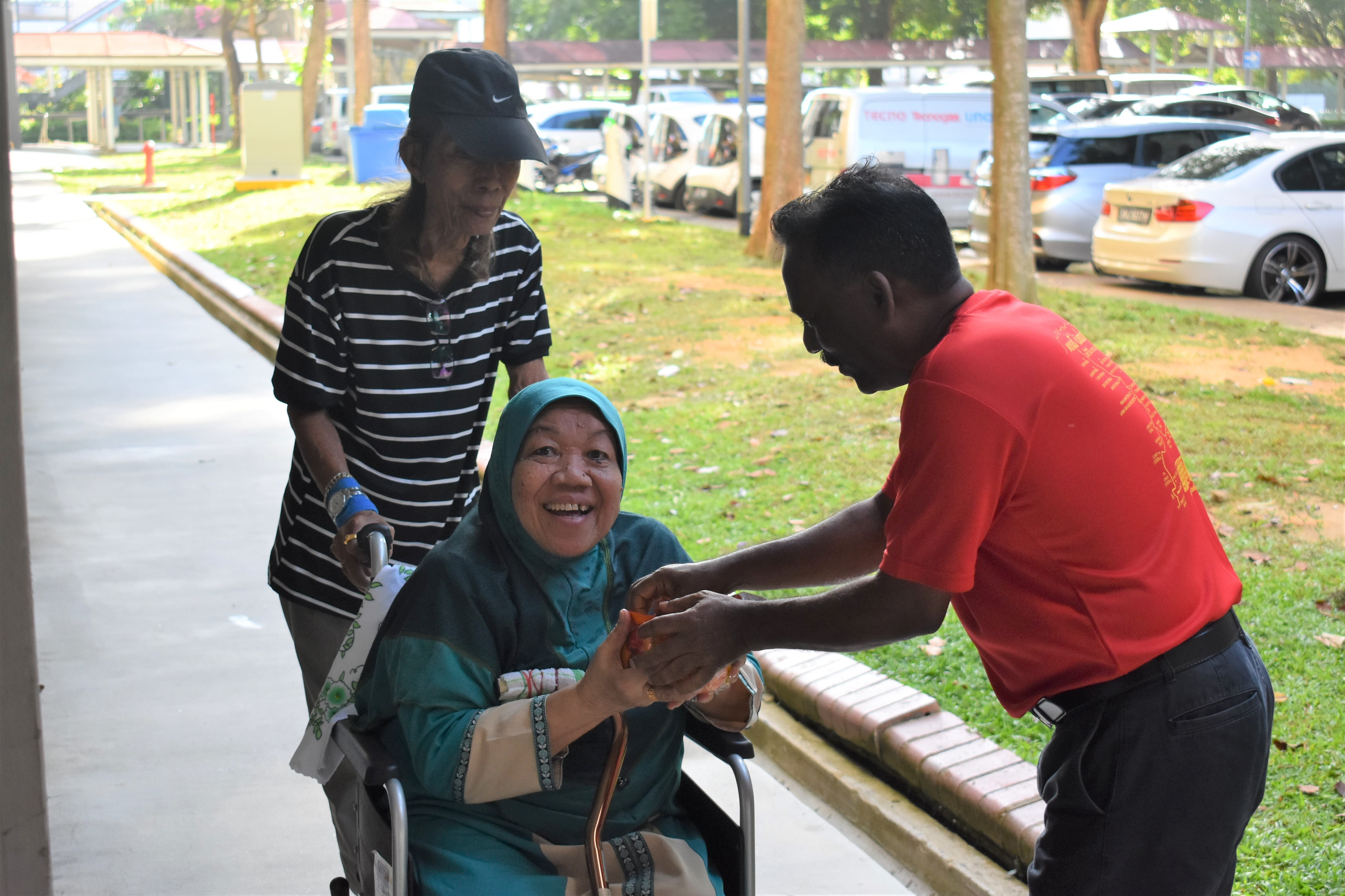 A smiling elderly woman in a teal hijab sits in a wheelchair while a man in red shakes her hand outdoors.